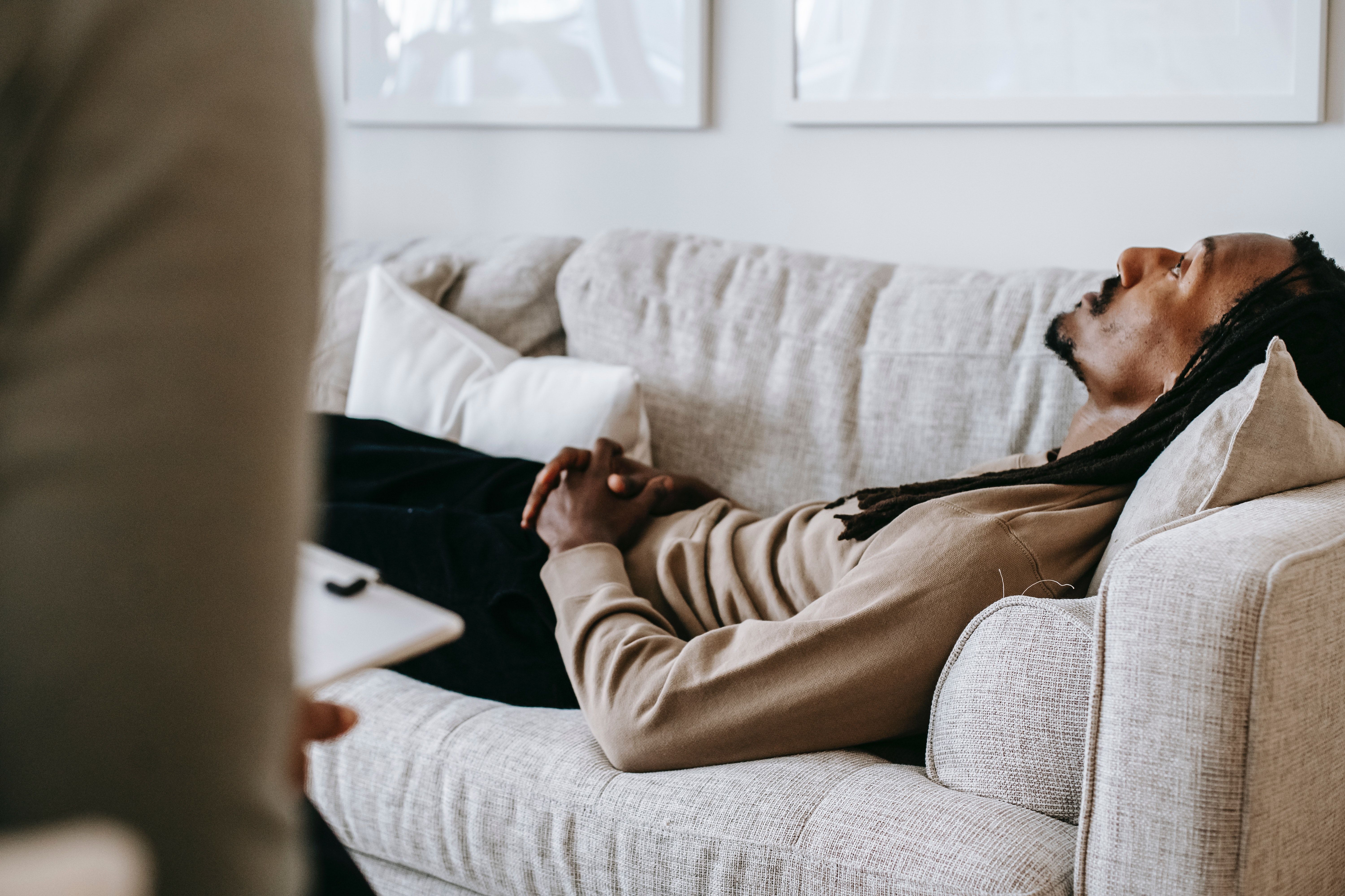 A black man with dreadlocks lies, despondent, on a gray loveseat before a therapist holding a clipboard.