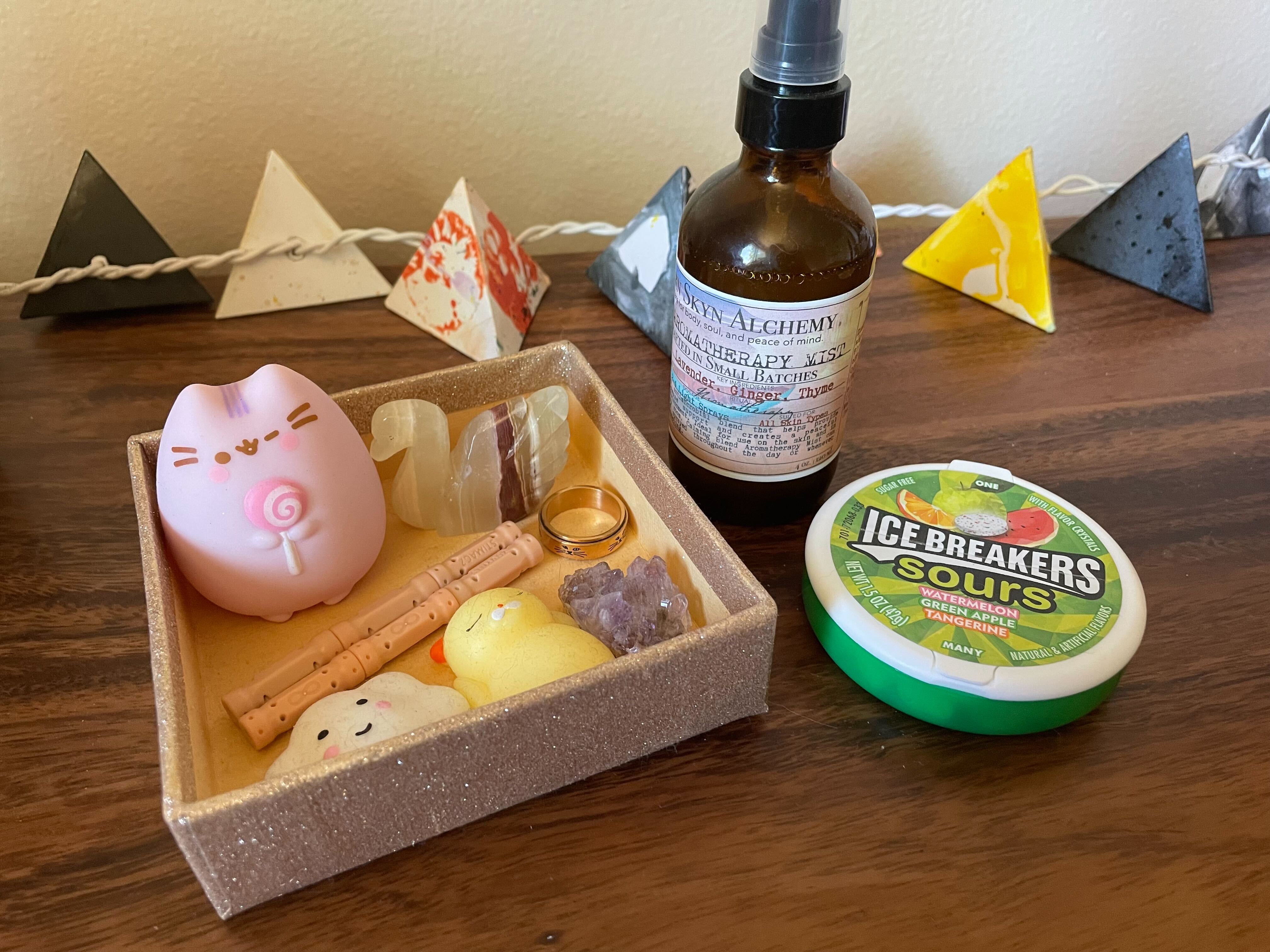 A small tray of fidget toys, including a pink cat, orange magnets, a purple crystal, and a rose gold spinning ring, as well as a spray bottle of essential oils and a green tin of sour candies. There are small paper lanterns lining the background.