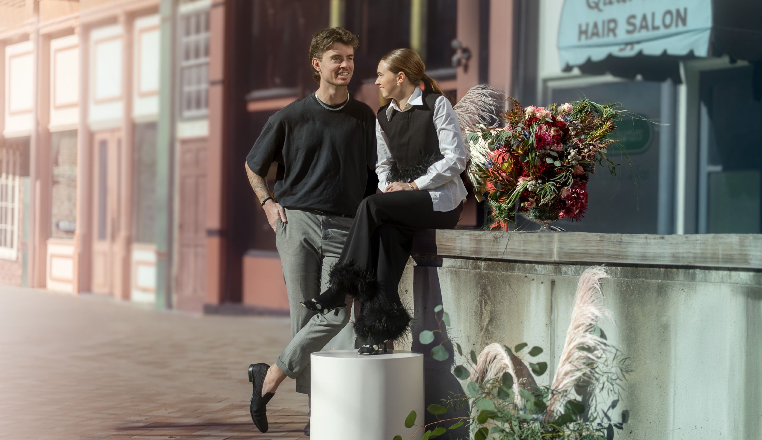 Couple on a downtown sidewalk outside a hair salon — one seated on a planter wall beside an arranged bouquet