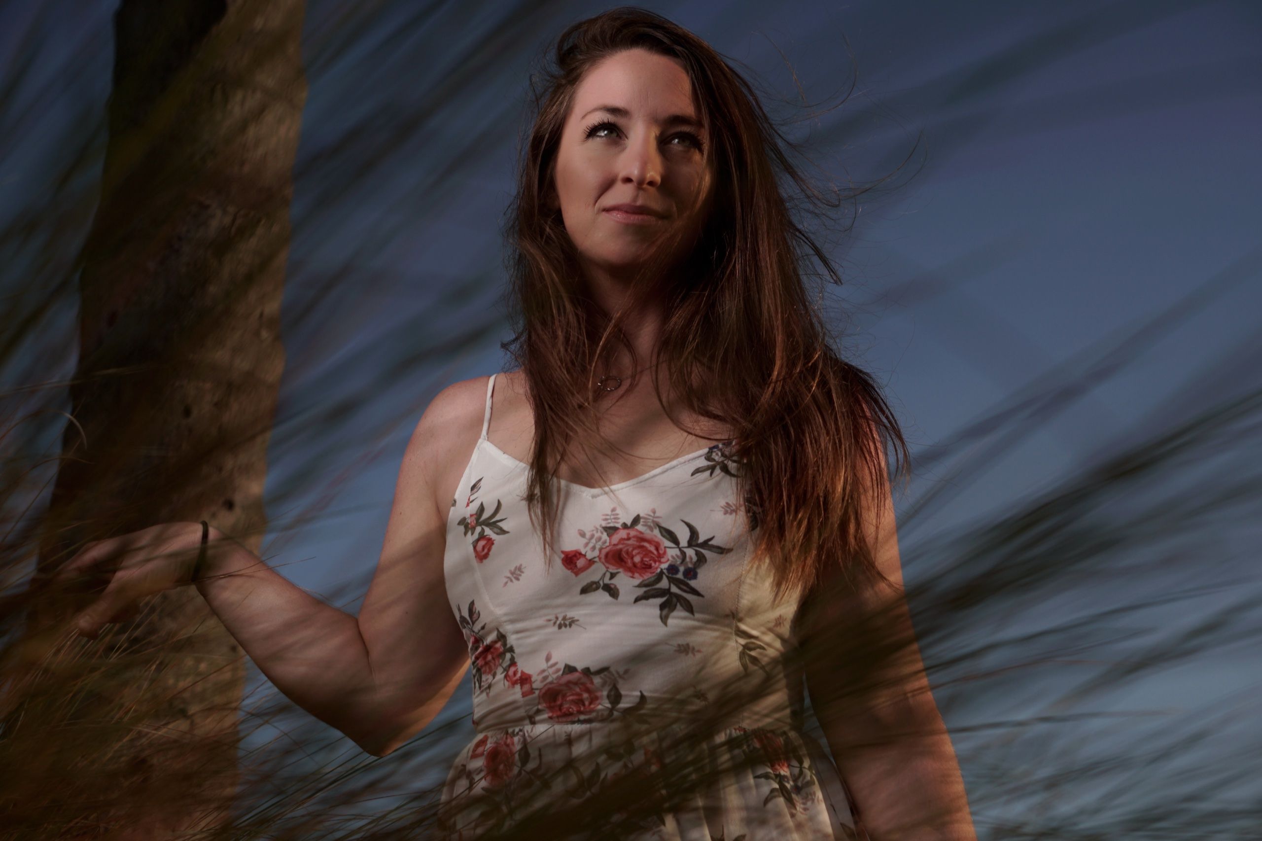 Portrait in a floral dress seen through wind-blurred grasses, dusk sky behind