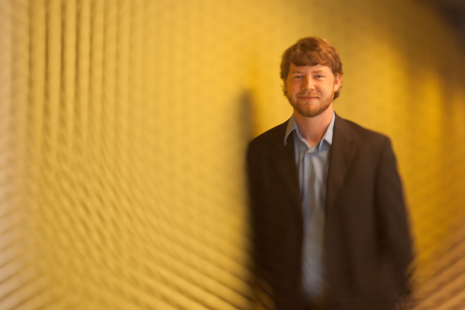A man in a blazer against a gold patterned wall, soft focus falling off around him