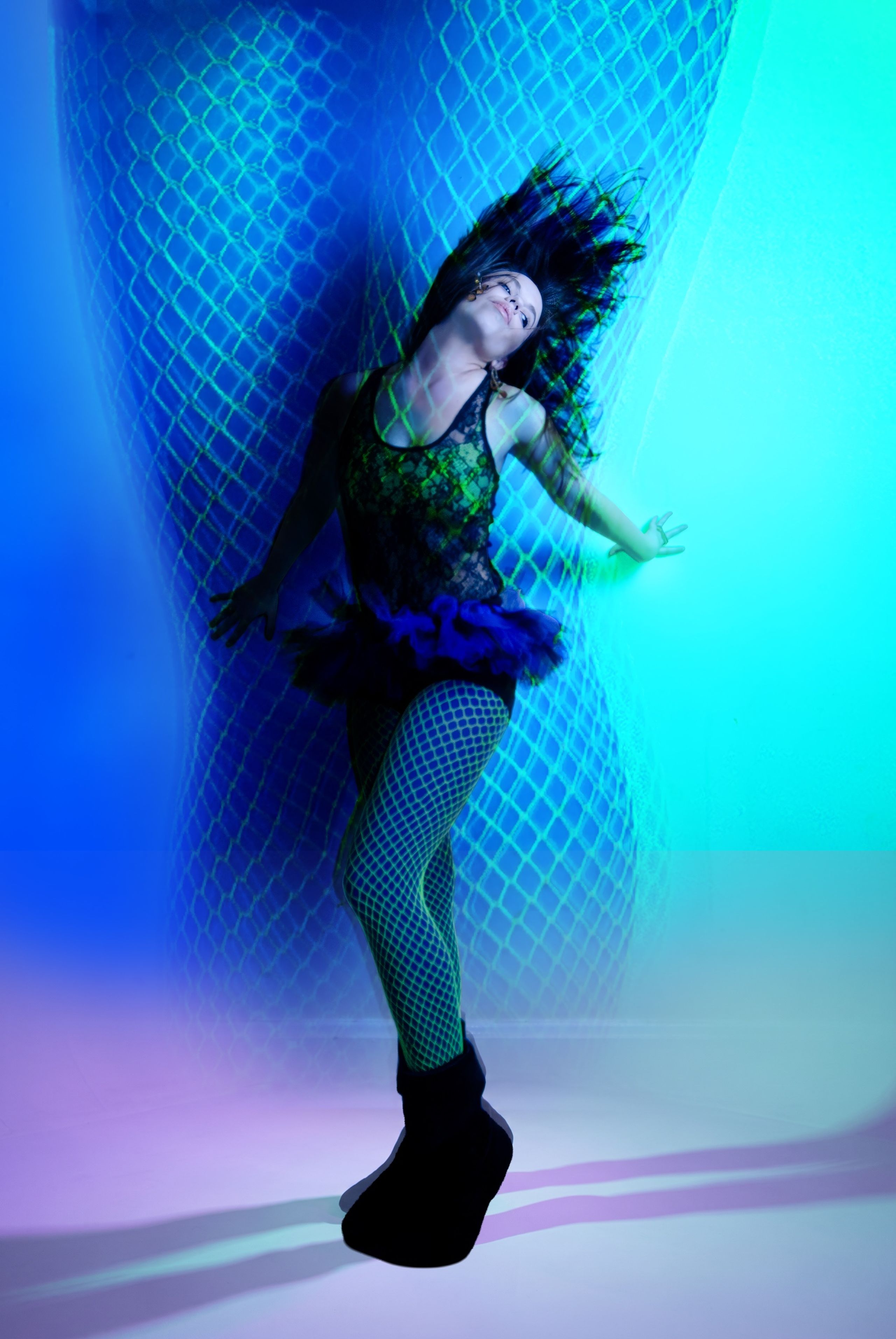 A dancer arches backward in blue and green gelled light against a patterned backdrop, in a tulle skirt and fishnets.