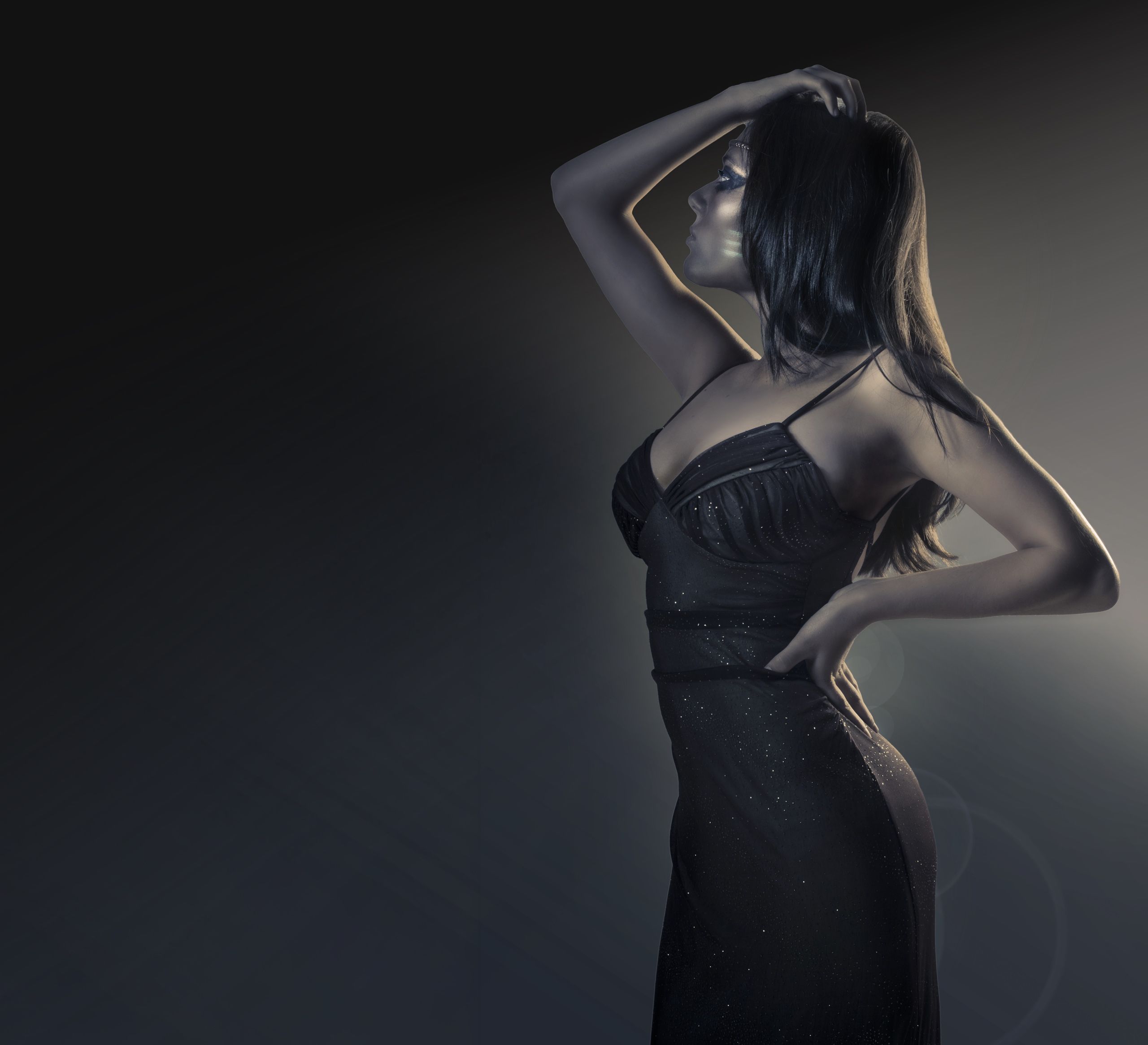A woman in a beaded black gown turns away from the camera with one arm raised, lit low against a graduated studio ground.