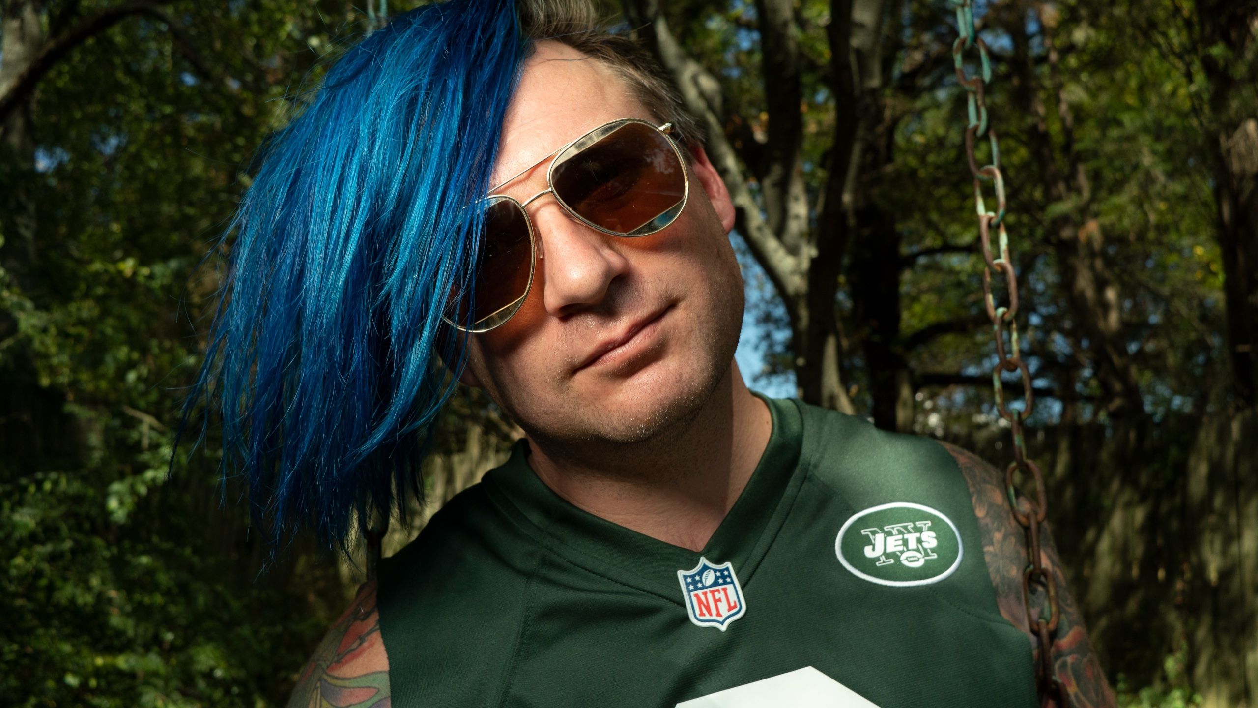 Promo portrait — blue-haired man in aviators and a New York Jets jersey under mossy oaks