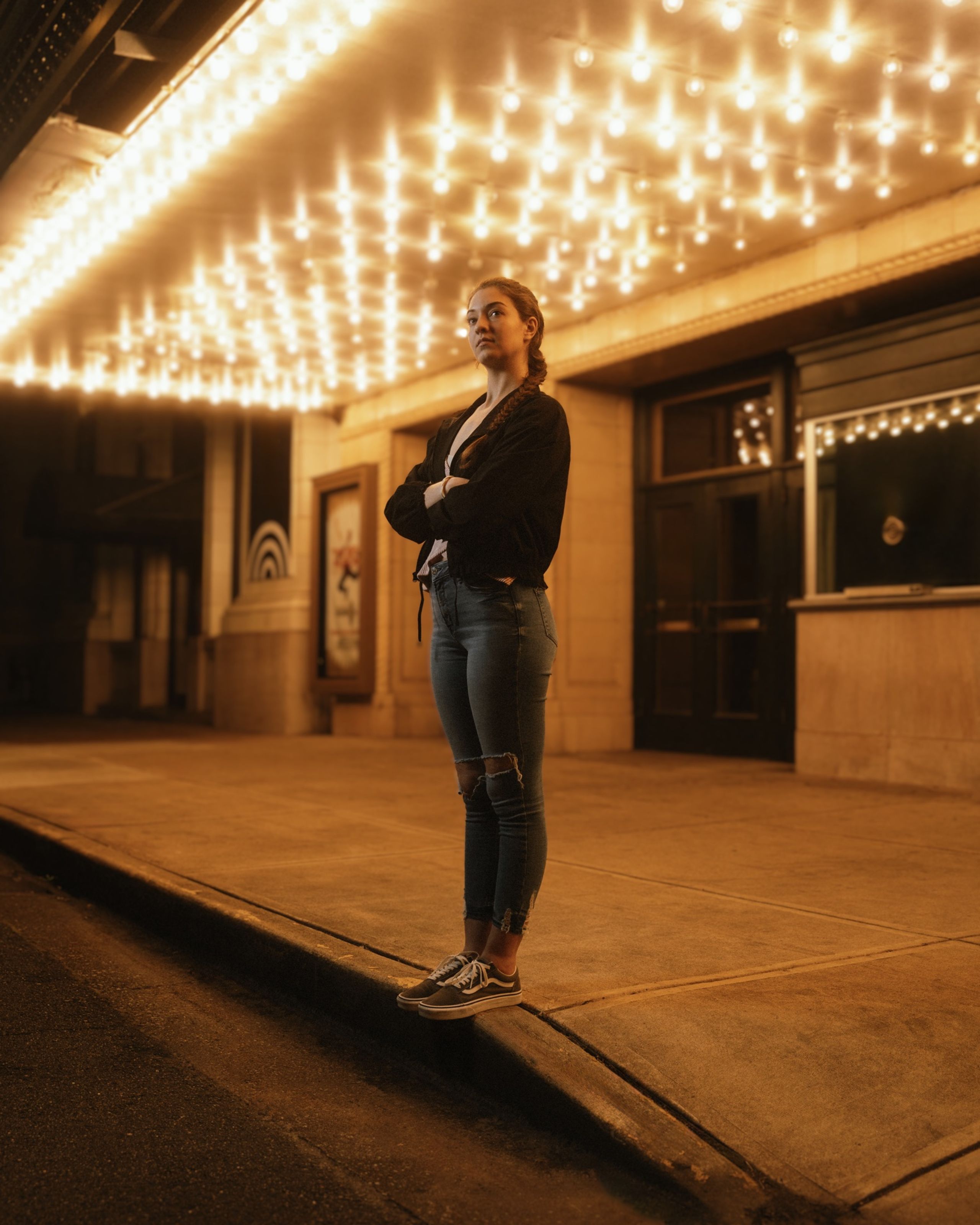 Hannah standing on the curb under a downtown theater marquee at night, arms crossed