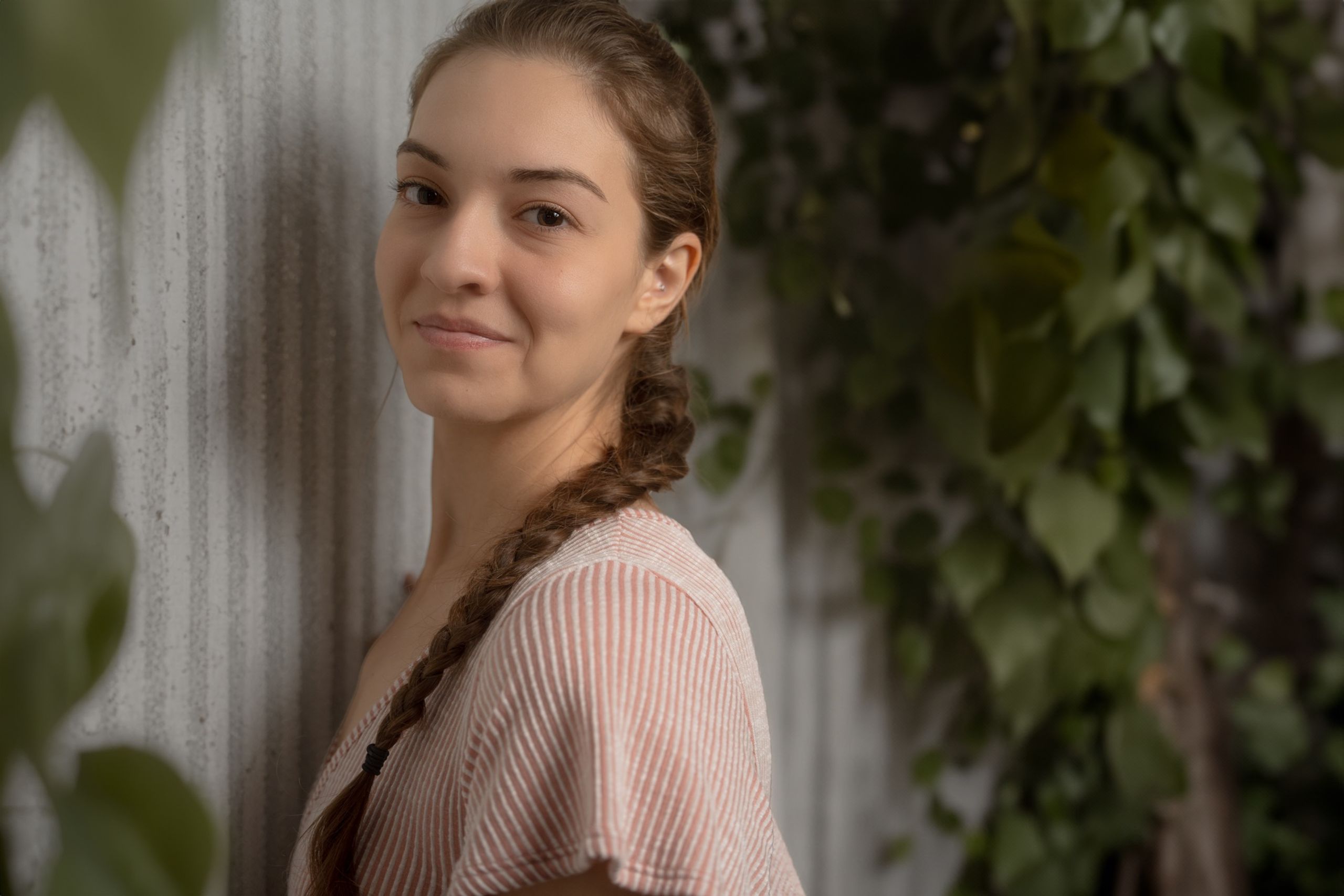 Hannah leaning against a corrugated wall, long braid over one shoulder, framed through soft foliage