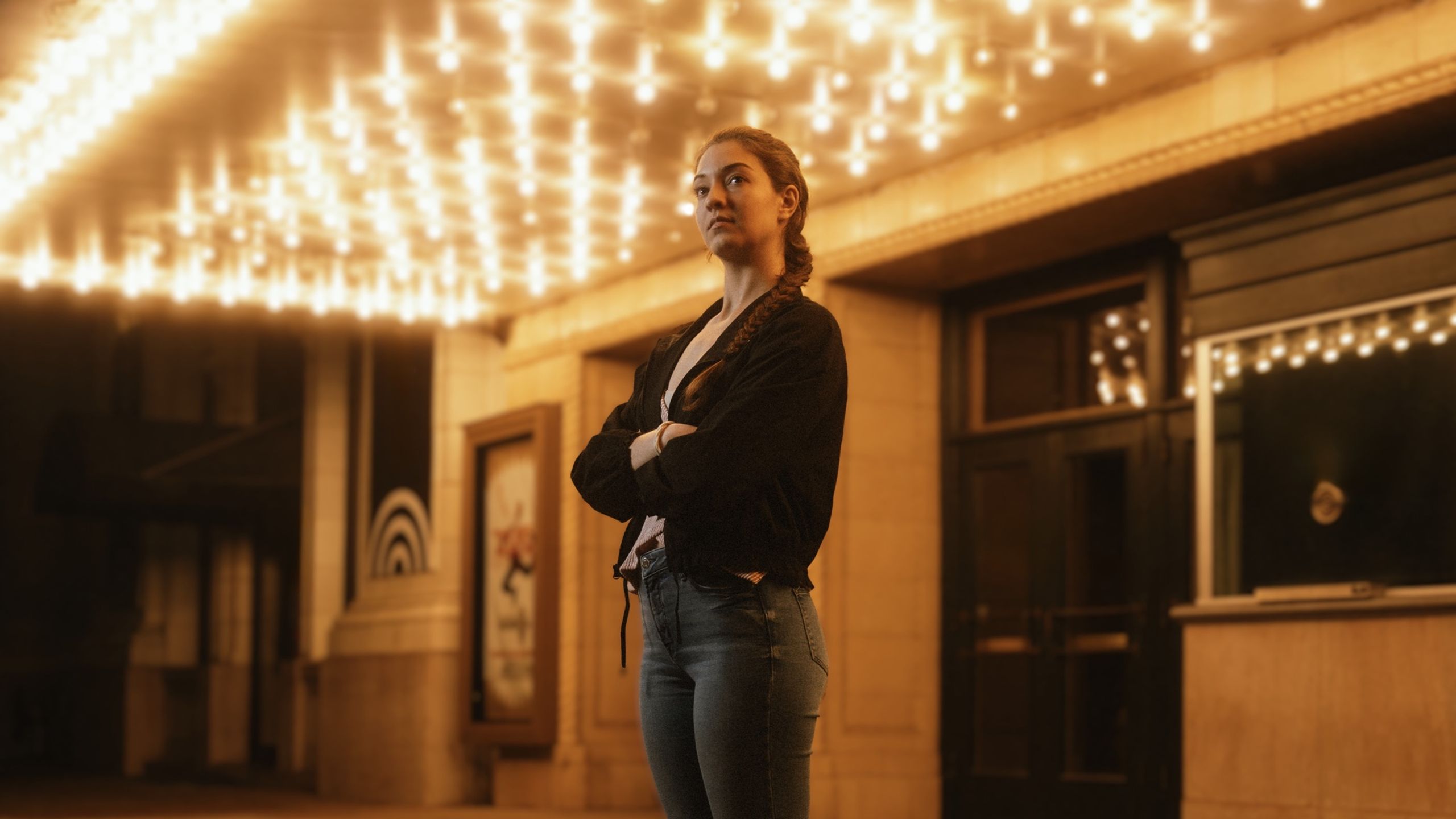 Hannah under a theater marquee at night, arms crossed beneath a field of warm bulbs