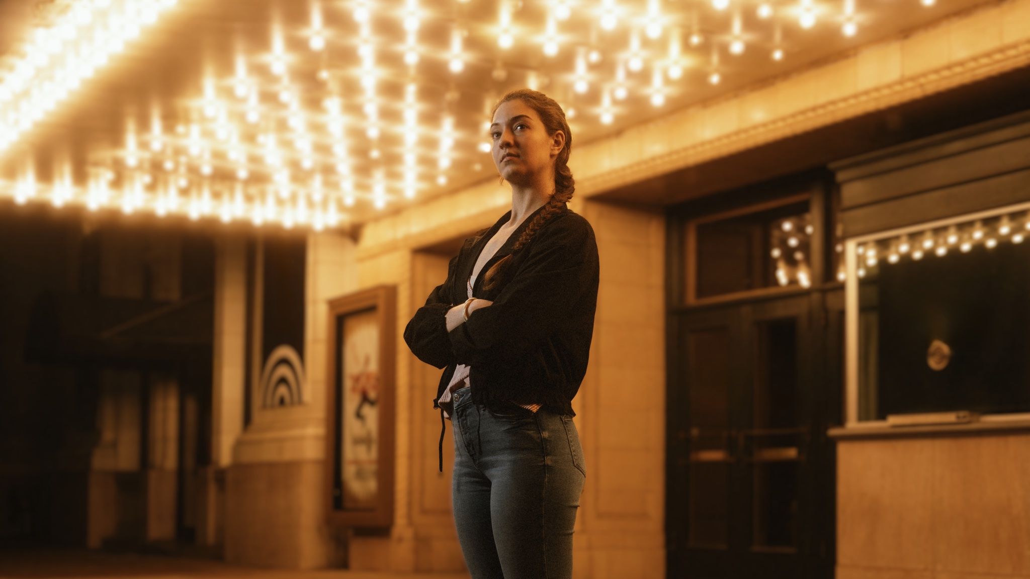 Hannah under a theater marquee at night, arms crossed beneath a field of warm bulbs