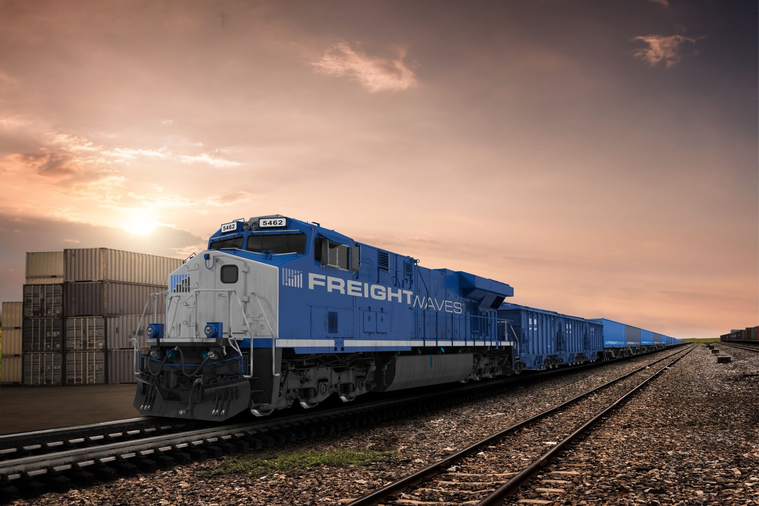 Freight locomotive in FreightWaves livery pulling blue hoppers past stacked containers at sunset