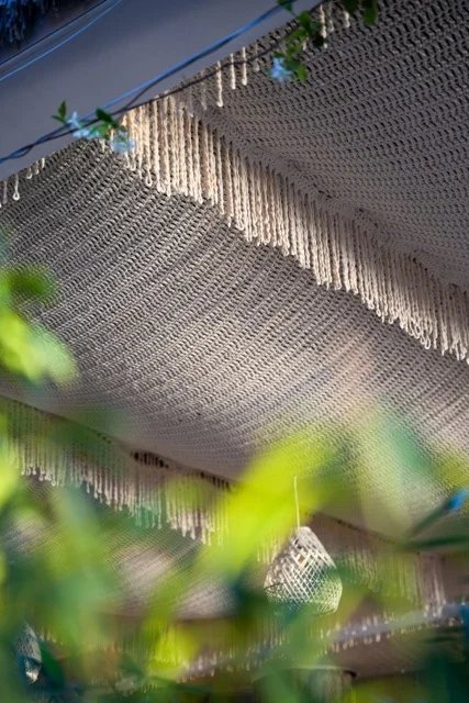 A cream open-knit canopy with a dense fringe edge above pendant lights, seen through foliage at So Beach.