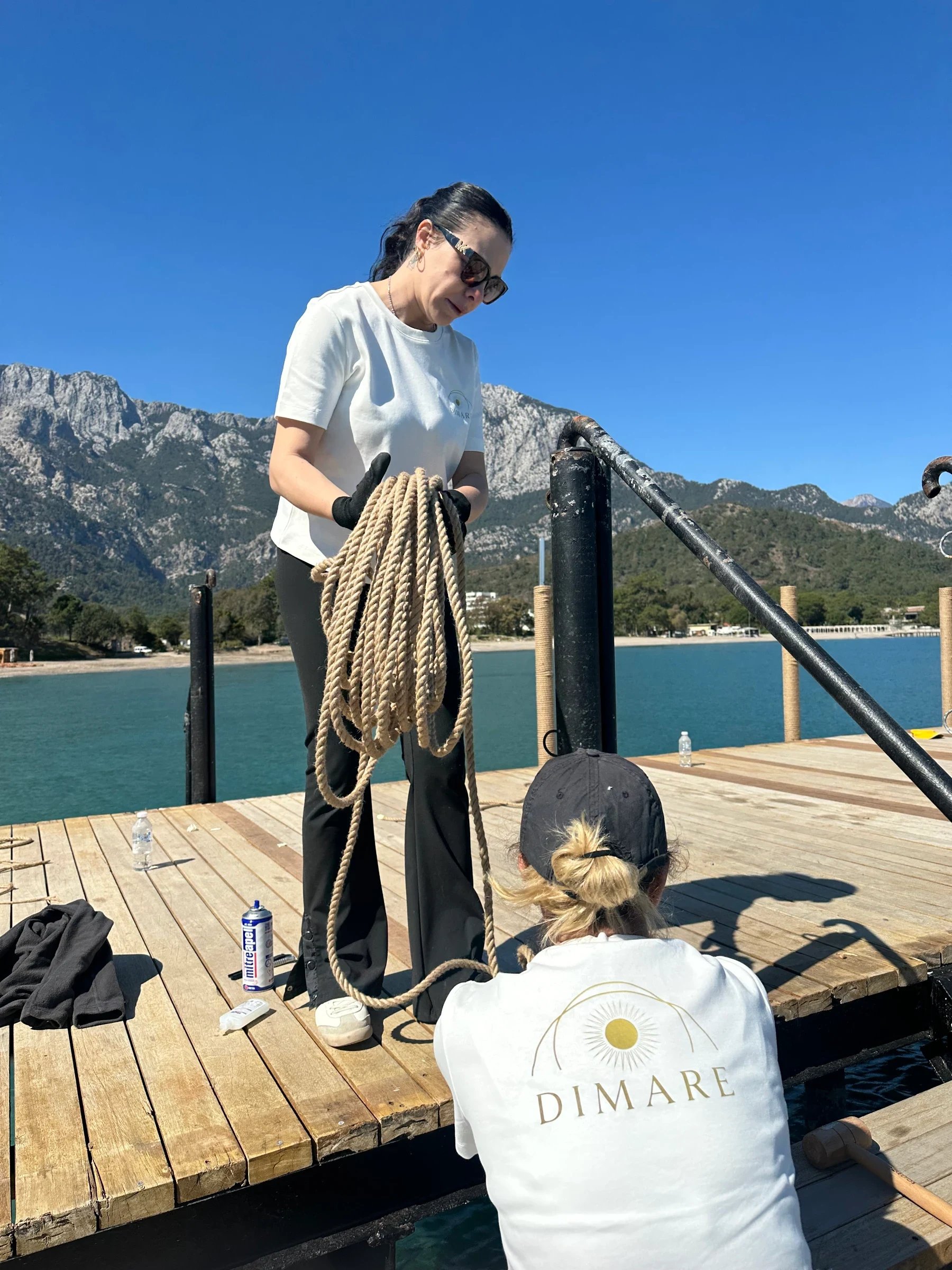 A person holding coiled rope beside a railing while another works at deck level.
