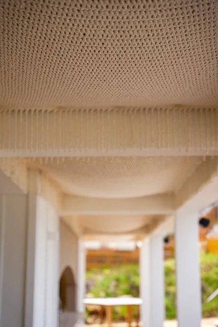 The underside of a cream knitted canopy with repeated fringed cross edges above a covered walkway at So Beach.