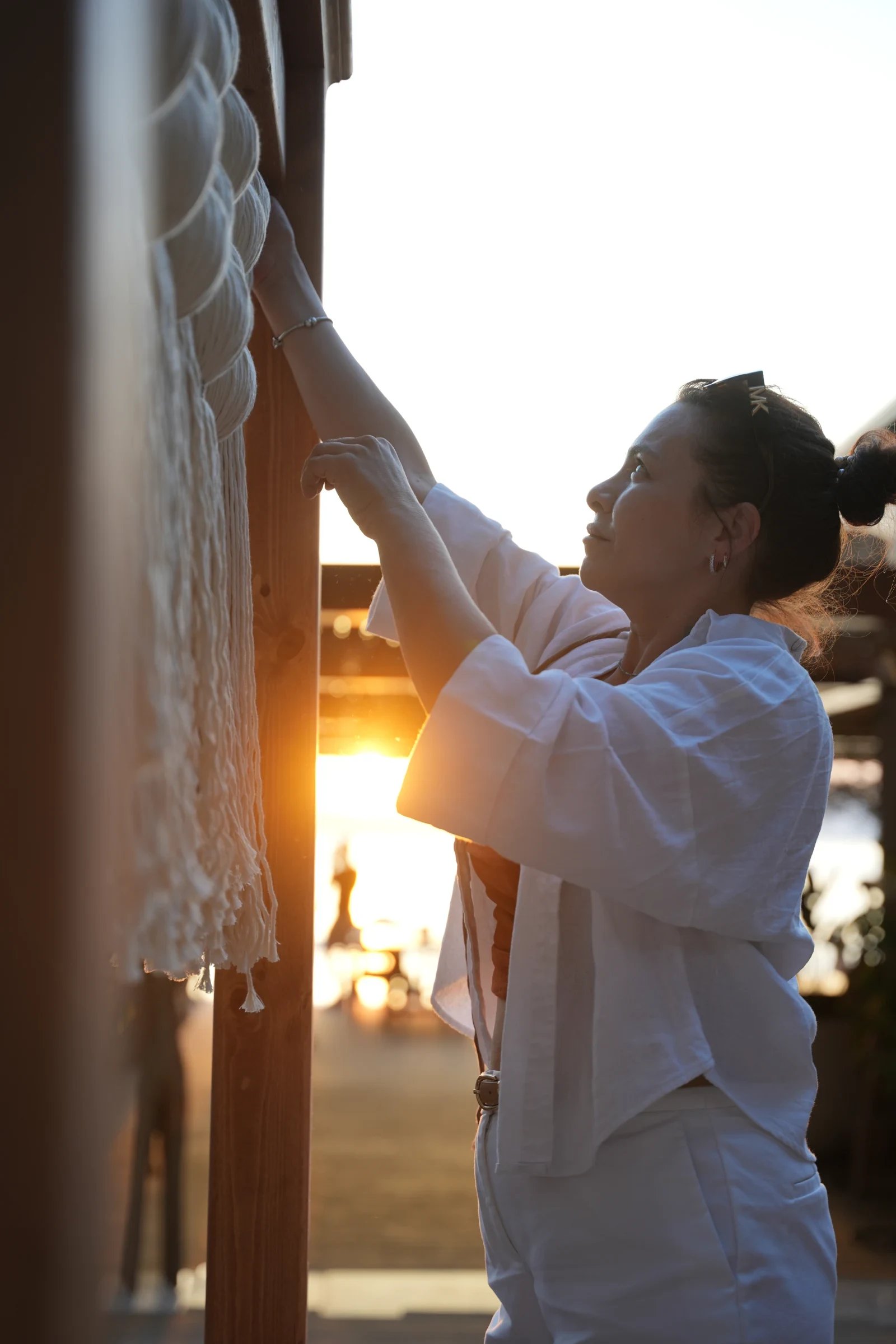 An artisan adjusting a cream fringed textile beside a timber opening in warm light.