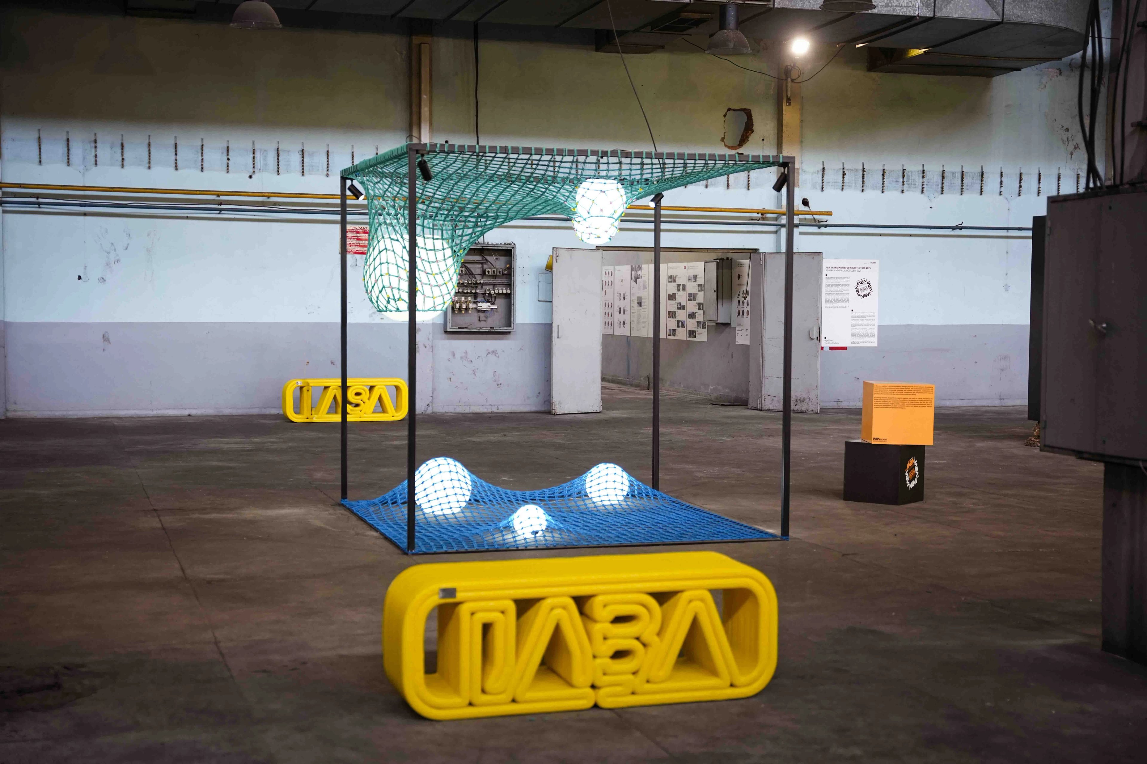 A framed teal-and-blue net installation with glowing spheres behind yellow biennial lettering at IABA 2025.