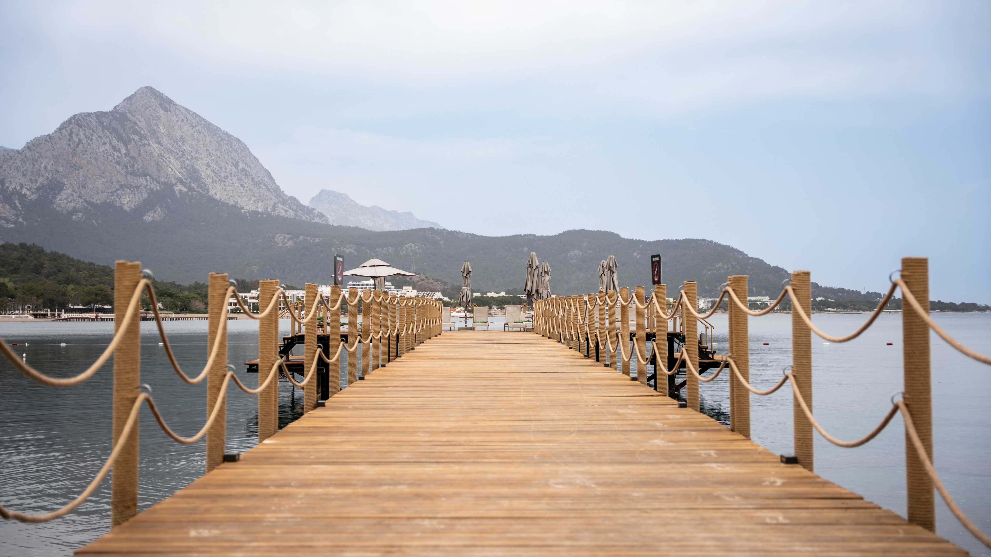 A timber jetty lined with rope-wrapped posts and looping rope rails, with mountains beyond at NG Phaselis Bay in 2025.