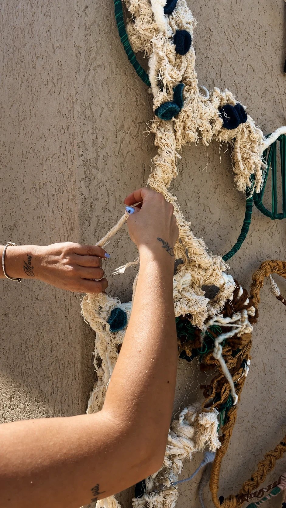 Hands arranging pale fibres on a textured wall composition with coloured rope details.