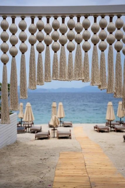 A cream sphere-and-tassel opening framing a timber path toward the sea and closed beach parasols at So Beach.