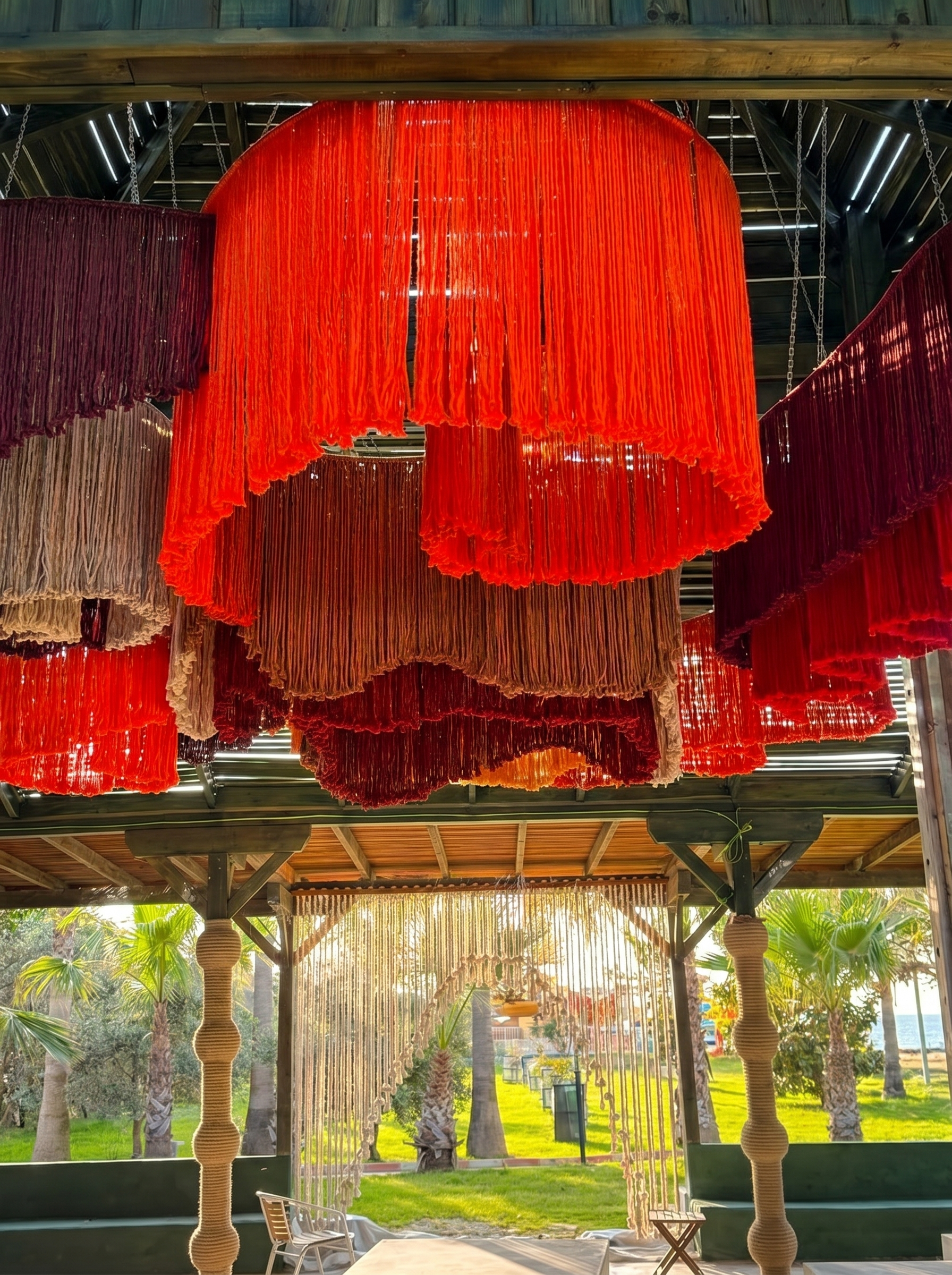 Layered red, burgundy and terracotta fringe forms suspended beneath a pavilion roof at MC Arancia Resort Hotel.