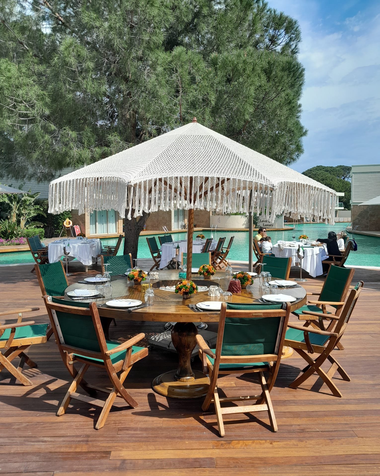 A cream fringed parasol above a set round dining table with timber chairs beside a pool at Rixos.