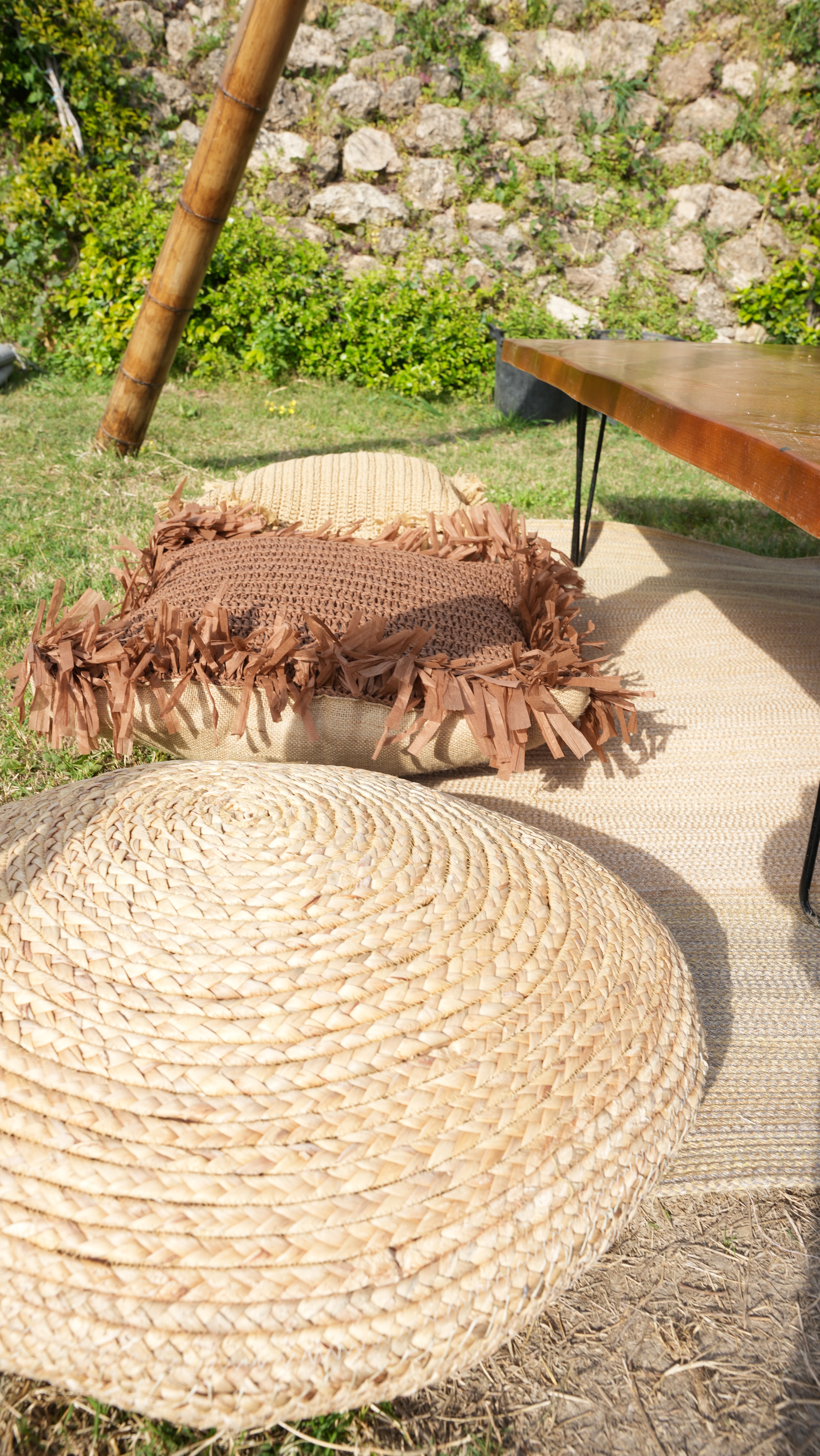 A round woven pouf and fringed floor cushion beside a garden table on the lawn at Lucky Monkey Hotel.