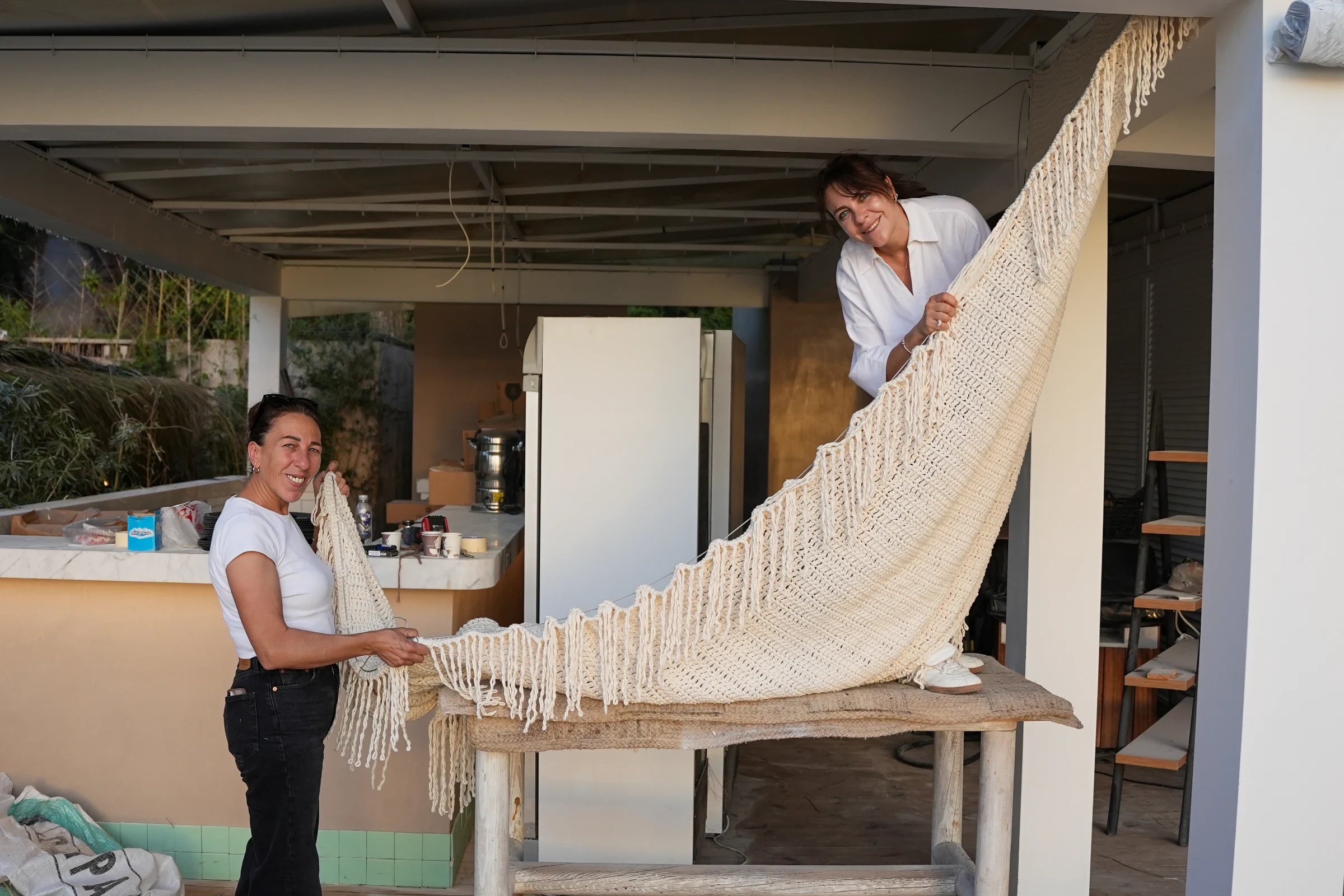An artisan lifting a large cream knotted textile with fringe over a worktable.