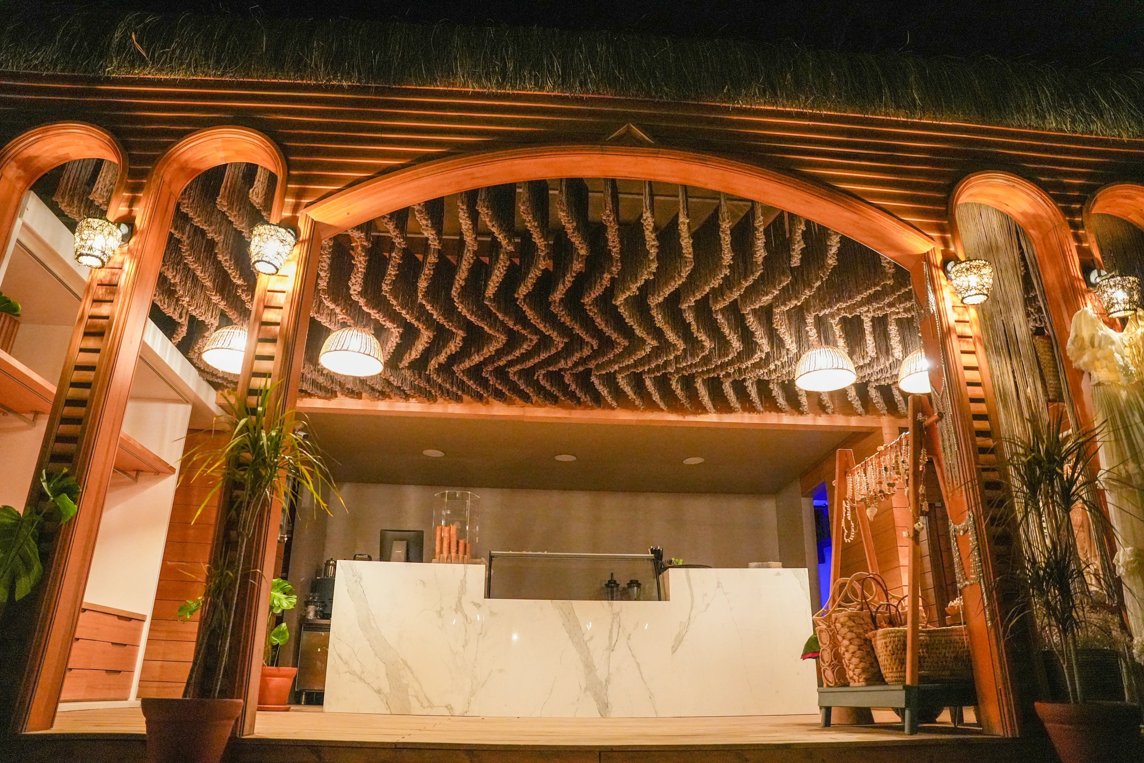 A dark chevron-patterned fringe ceiling above an illuminated bar with arched timber framing at So Beach.