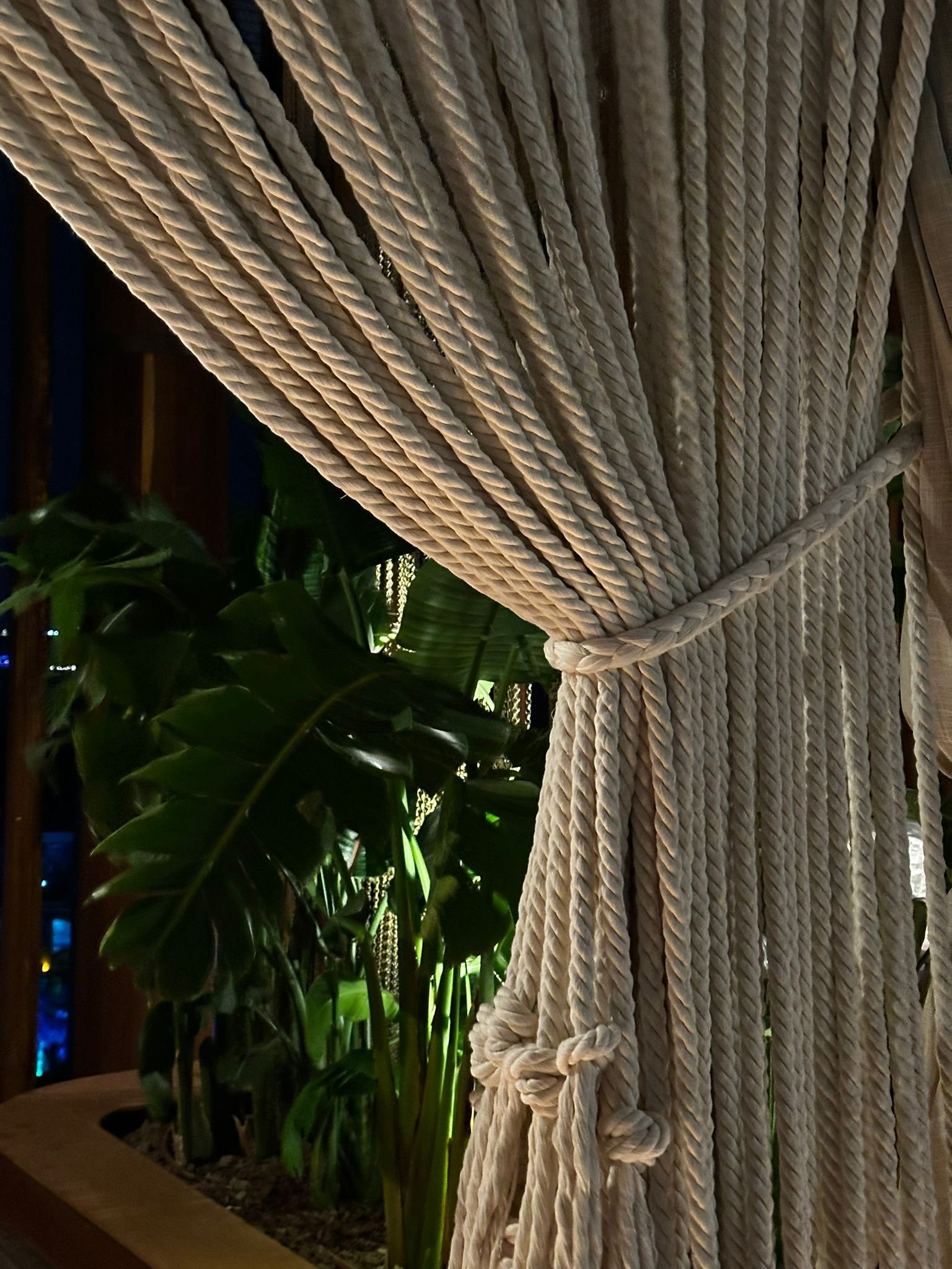 Close view of twisted cream curtain strands gathered by a rope tieback beside green foliage at Regnum The Crown.