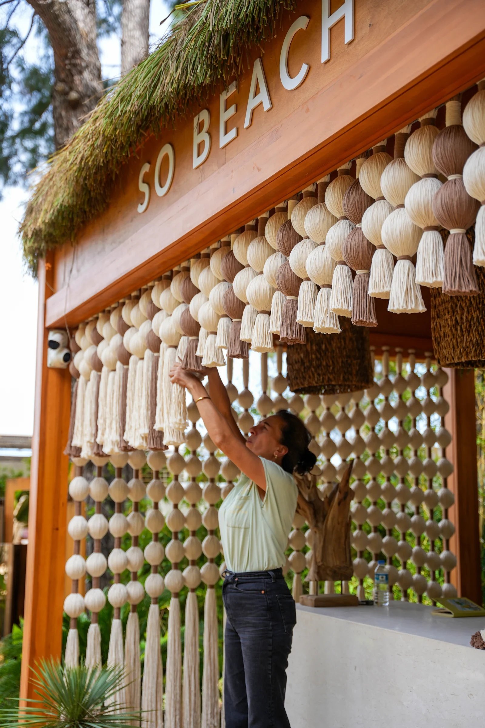 An artisan adjusting hanging cream woven forms and tassels beneath a timber fascia.