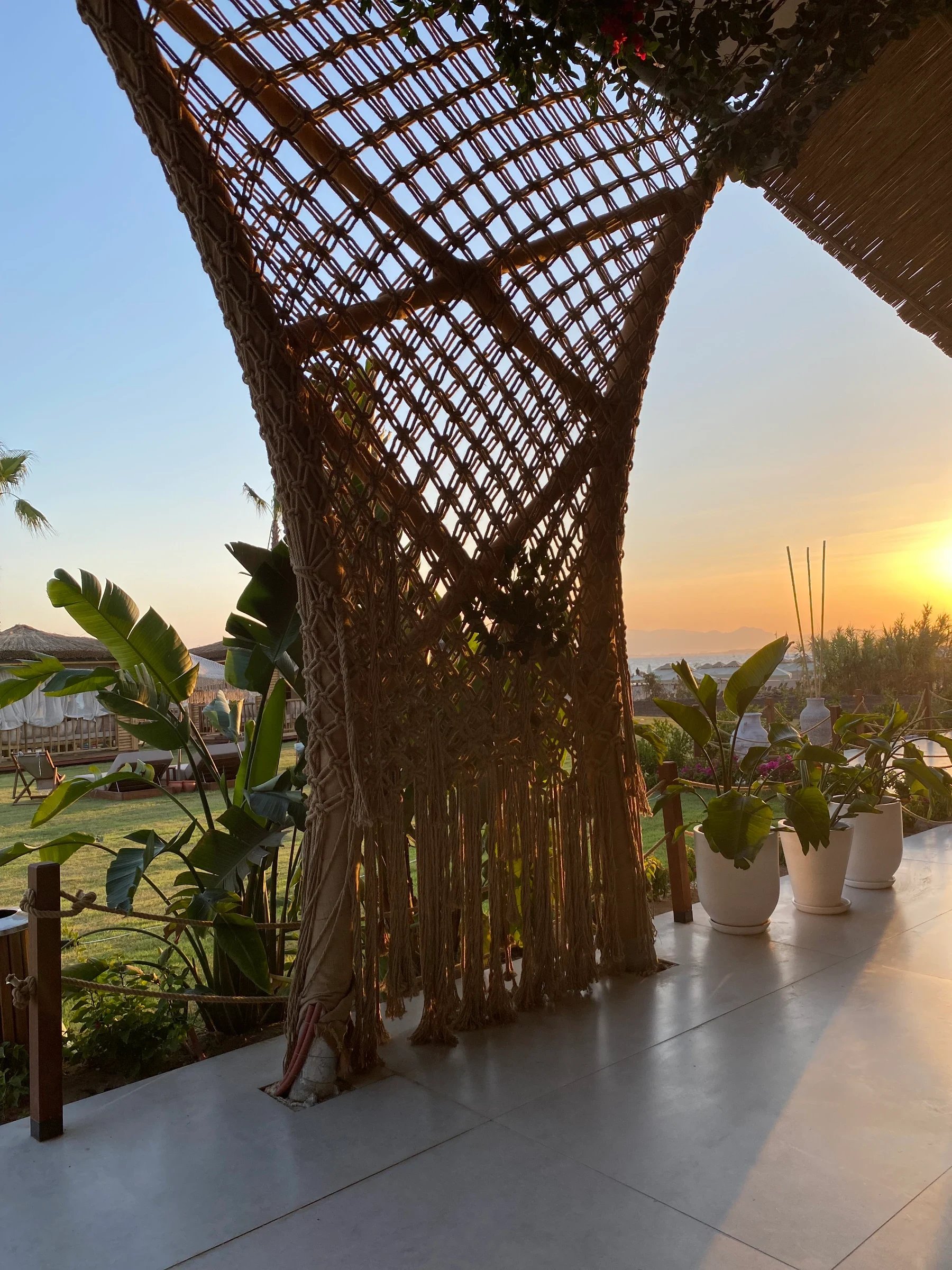 A curved open-knot rope screen with long fringe beneath an overhead frame at sunset at Ethno Hotels Belek.