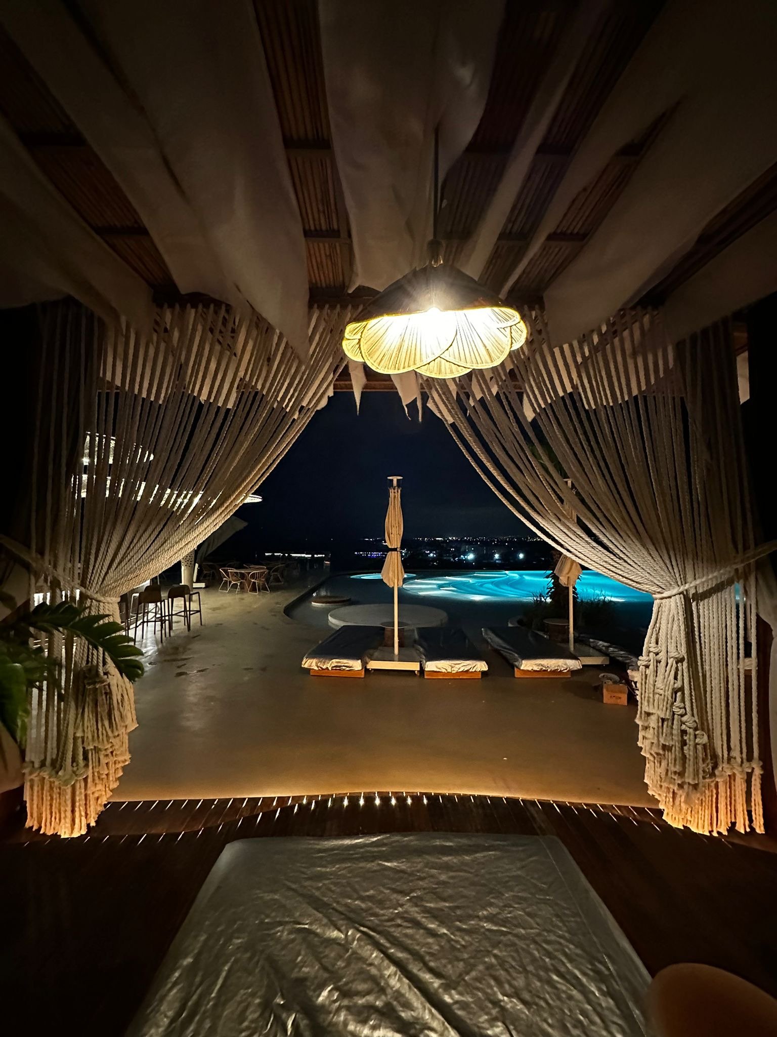 View from a cabana through tied-back cream rope curtains toward an illuminated pool at Regnum The Crown.