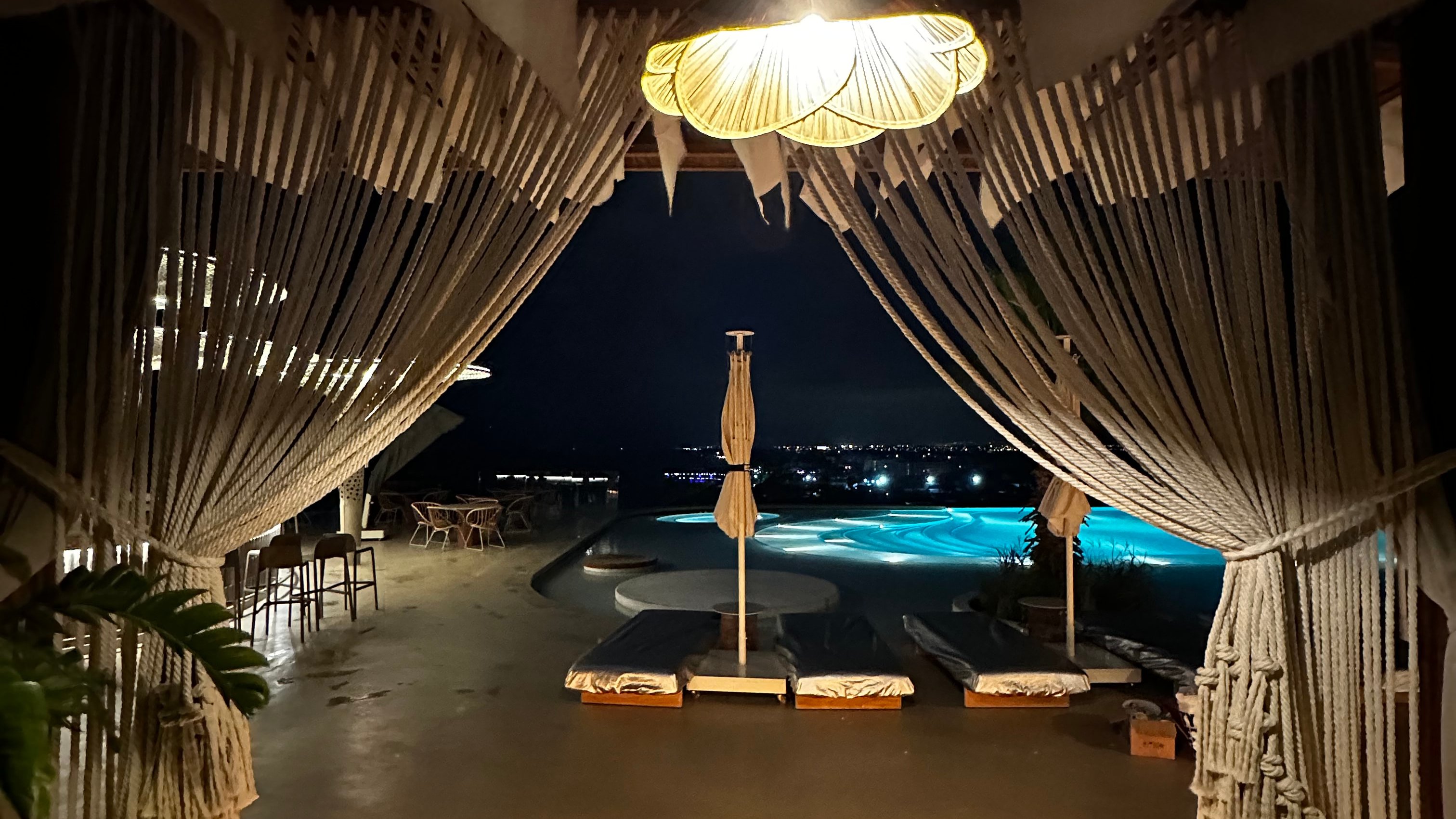 Cream rope curtains tied back beneath a lit cabana ceiling, framing a swimming pool at night at Regnum The Crown.