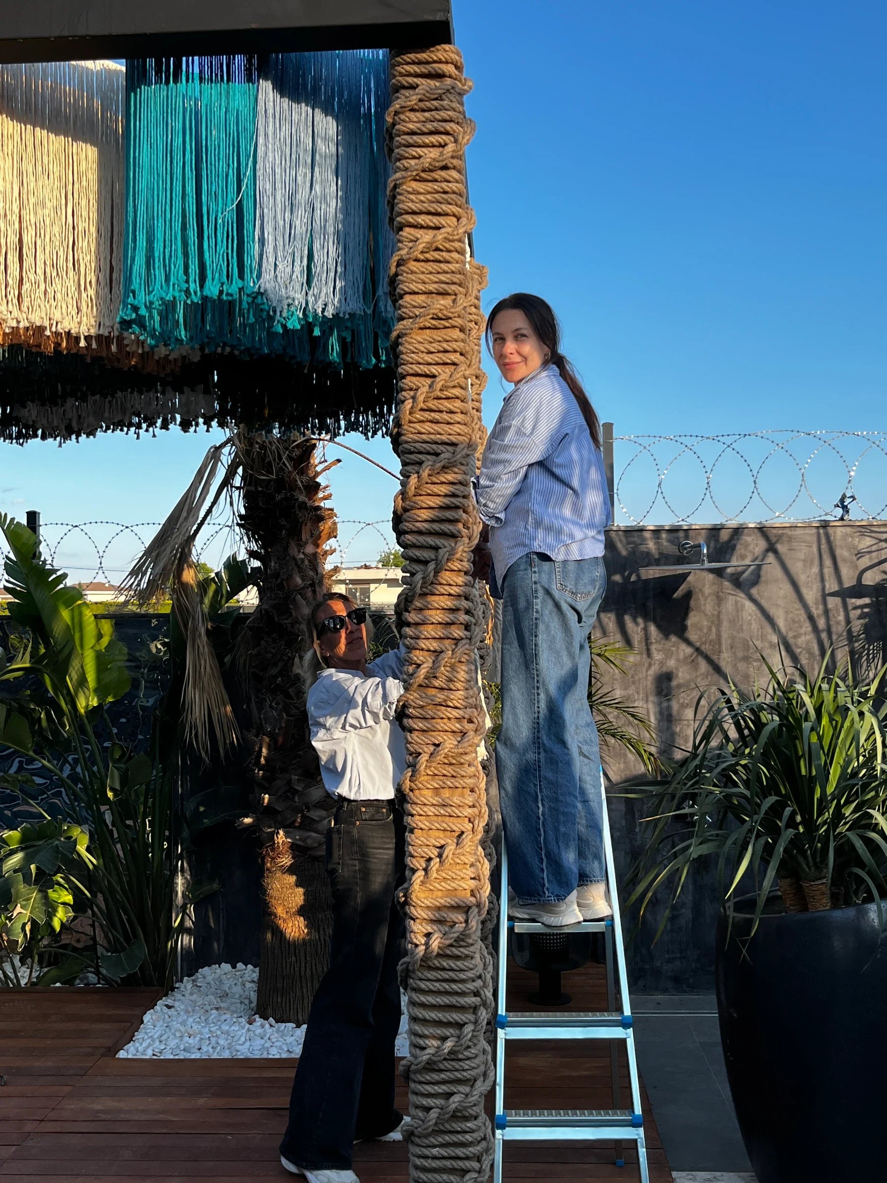 An artisan on a stepladder beside a rope-wrapped post and multicoloured fringe.
