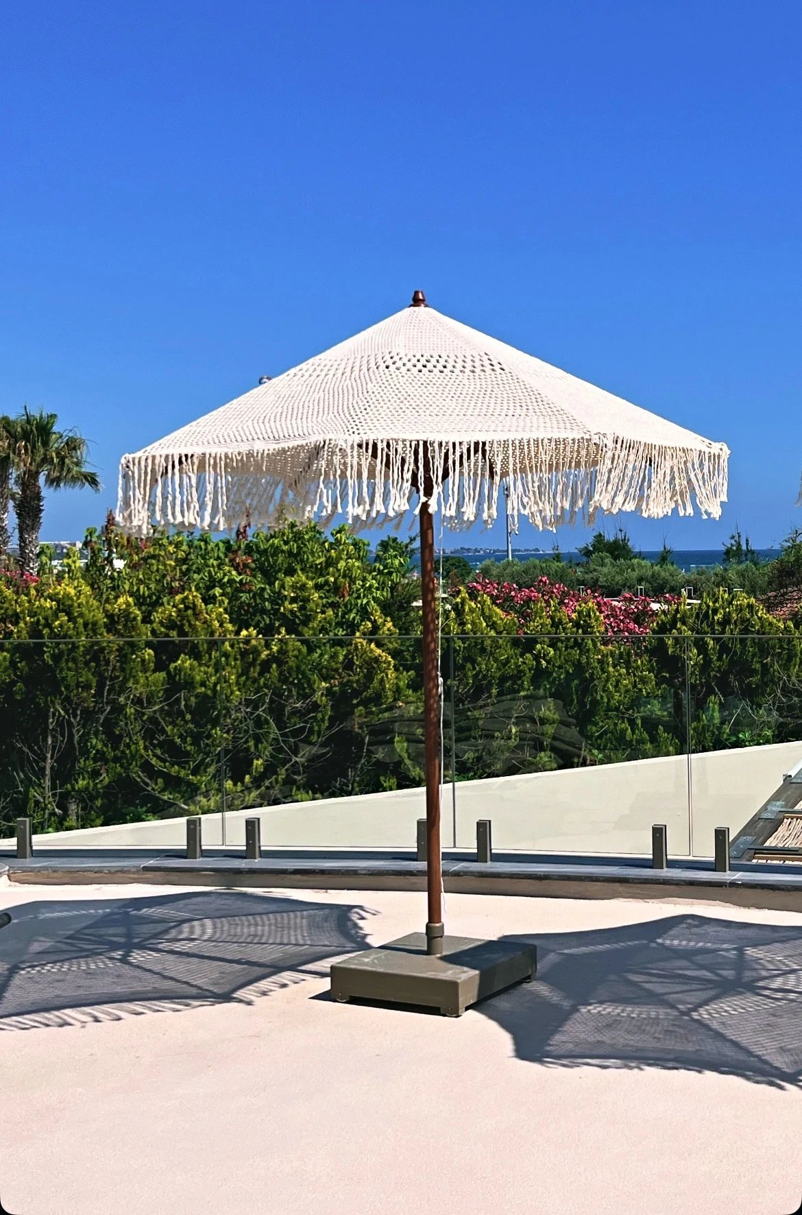 A single cream open-weave parasol with long fringe on a sunlit terrace above planting at Adalya Bliss.