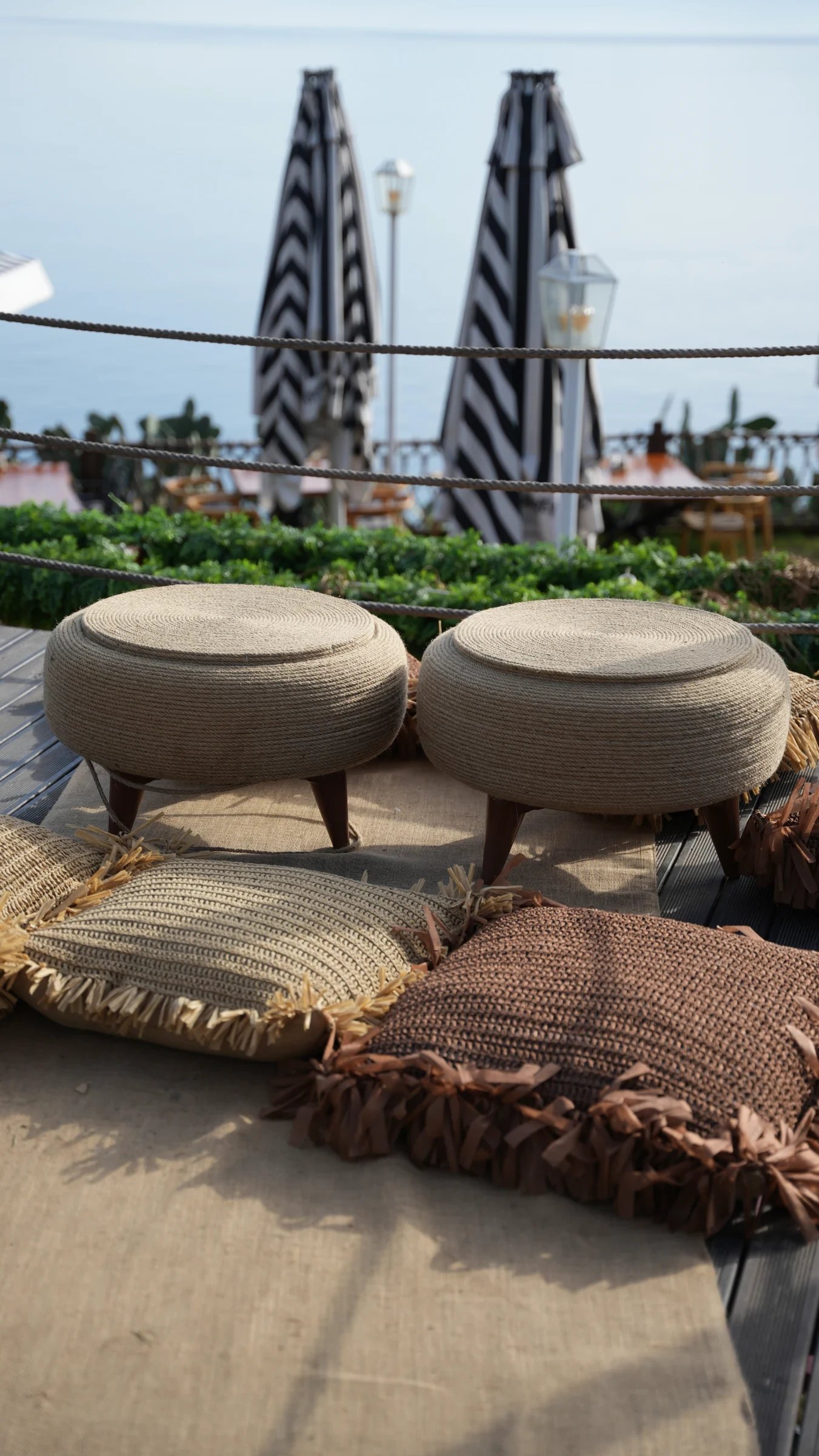 Two round woven poufs on short legs beside fringed cushions on a sea-view terrace at Lucky Monkey Hotel.