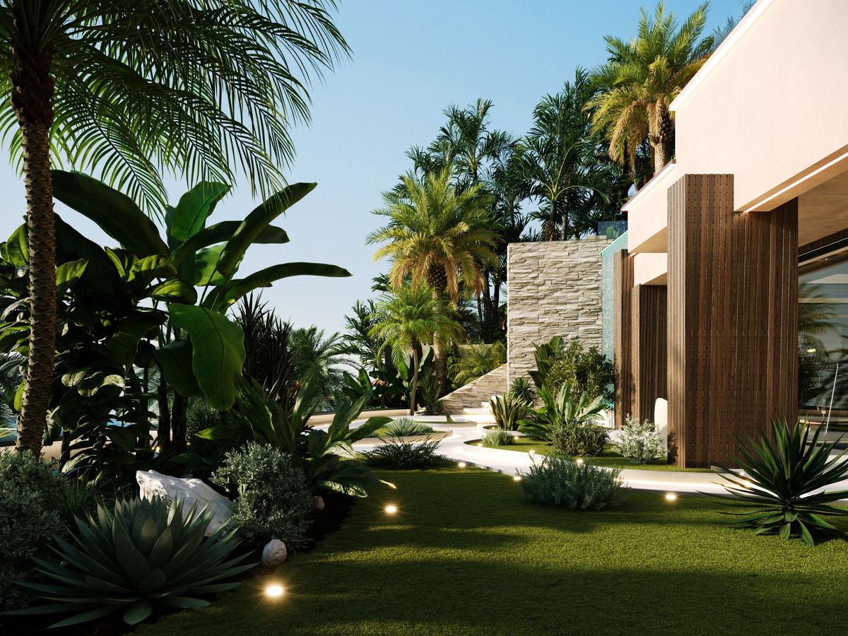 Jardín de una casa en la Costa del Sol al atardecer: palmeras, plataneras y agaves junto al césped, un muro de piedra y una fachada de listones de madera con luces bajas encendidas en el camino.