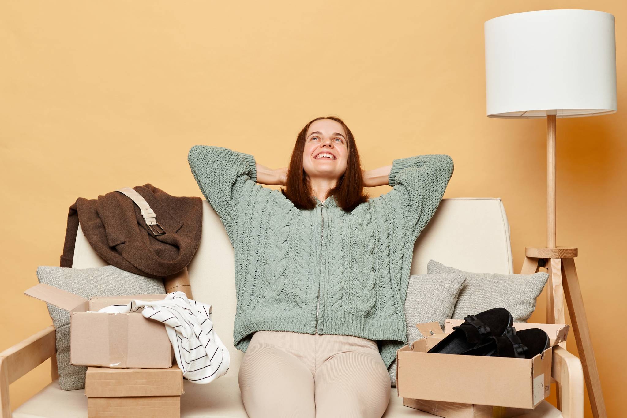 Woman sitting on couch looking relaxed with a pile of clothes and cardboard boxes around her.