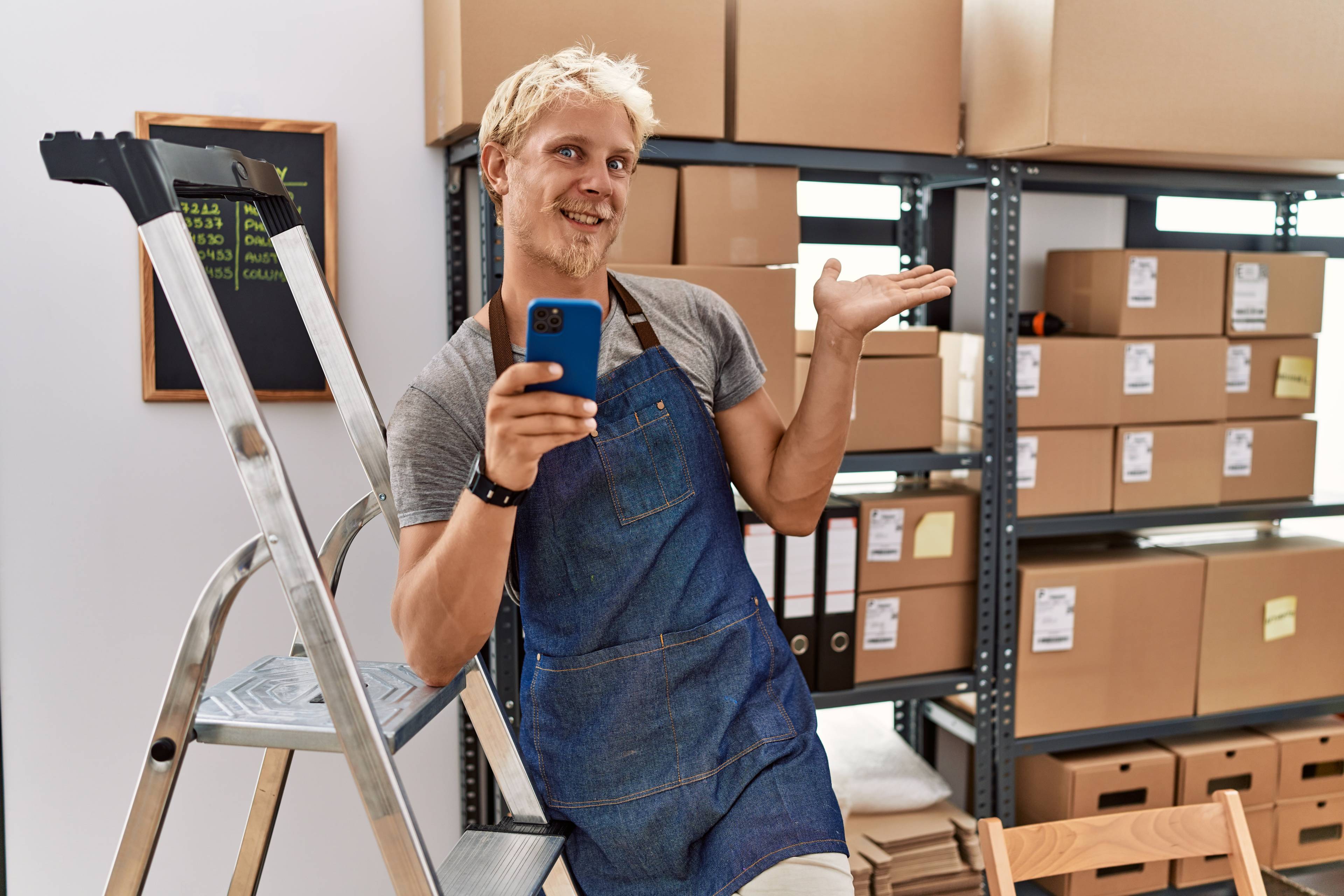 Young blond man using smartphone working at storehouse smiling