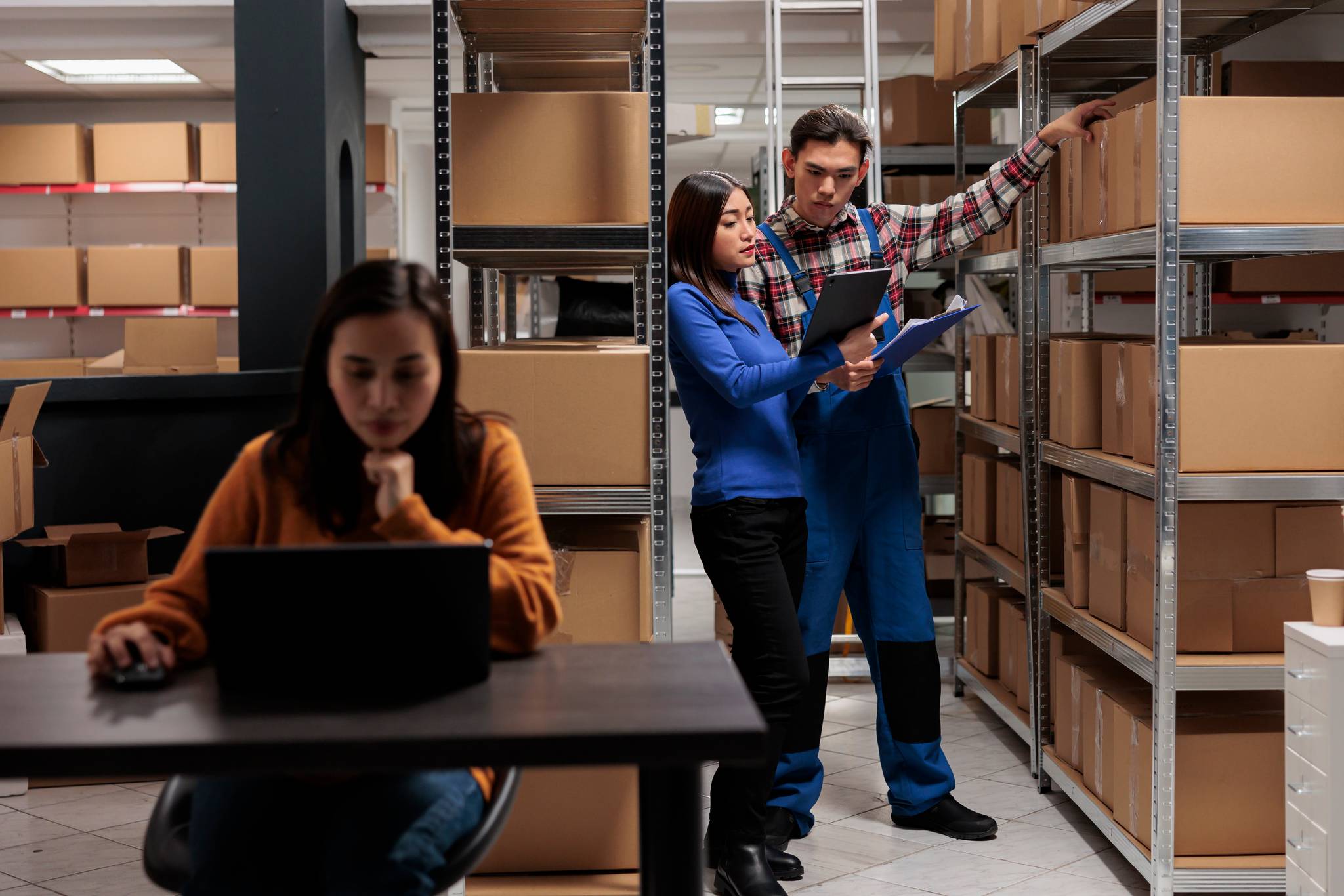 woman on laptop in warehouse with two people behind checking boxes