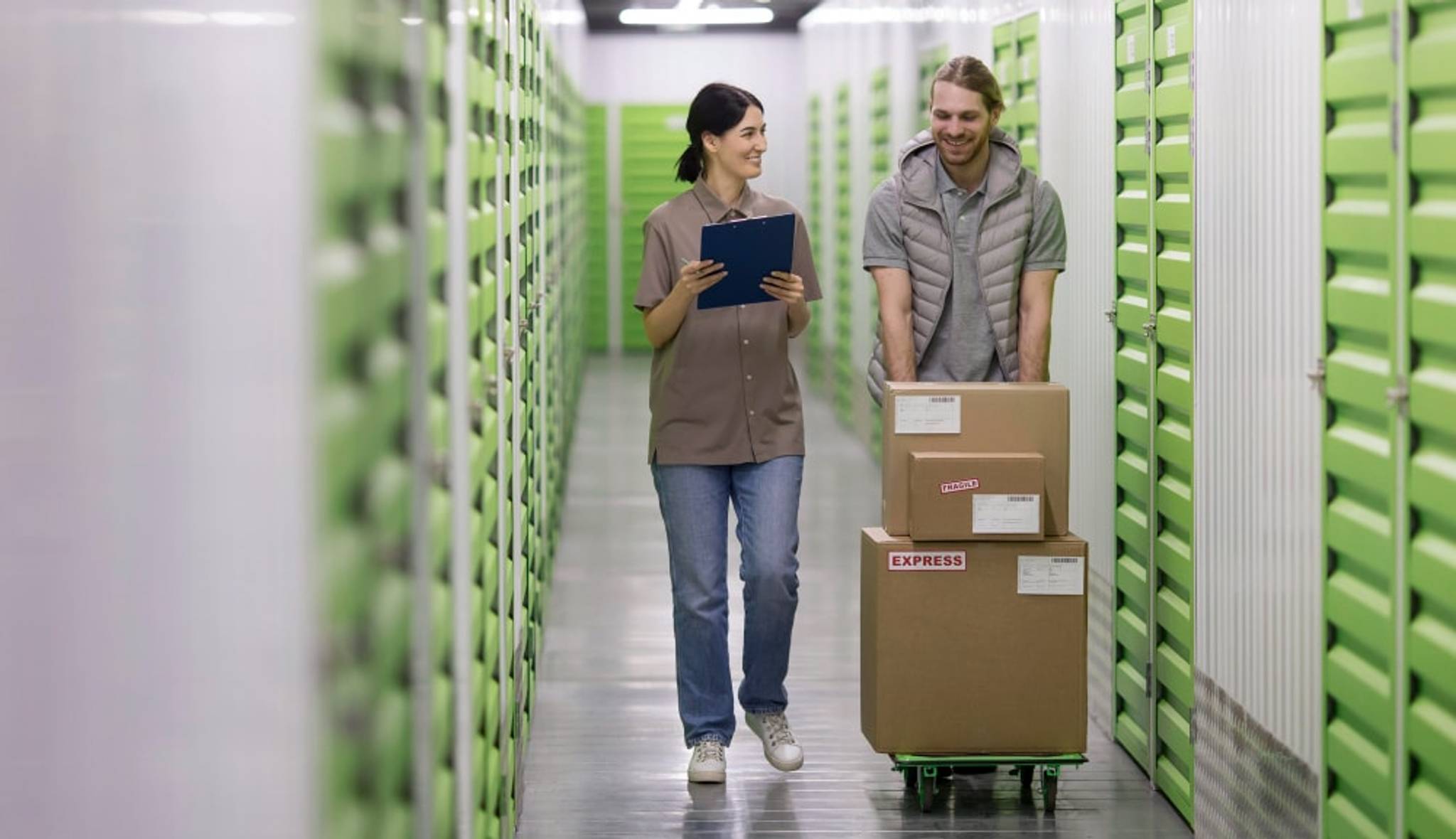 Two people walk down a hallway in a storage facility, carrying boxes