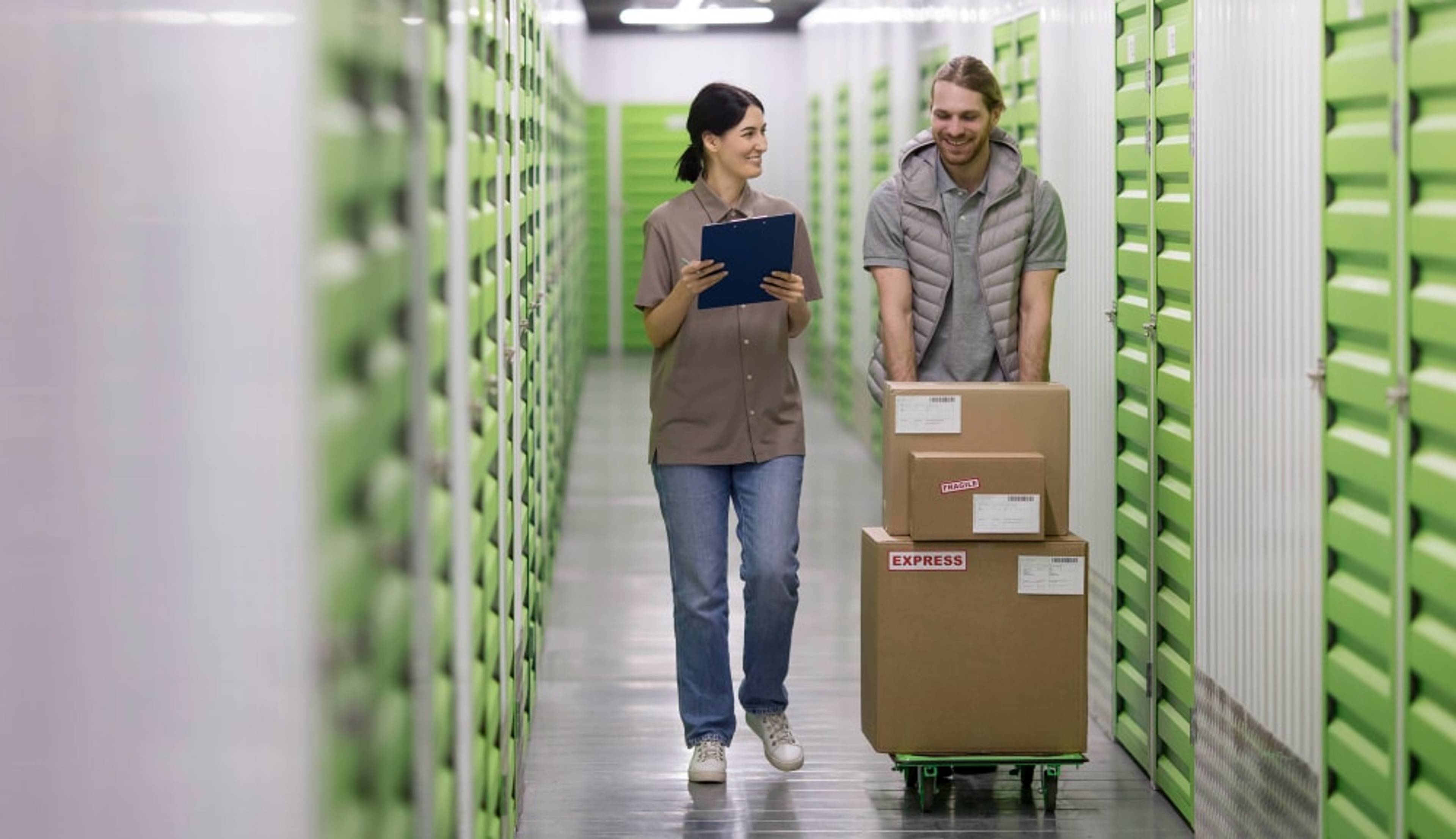 Two people walk down a hallway in a storage facility, carrying boxes