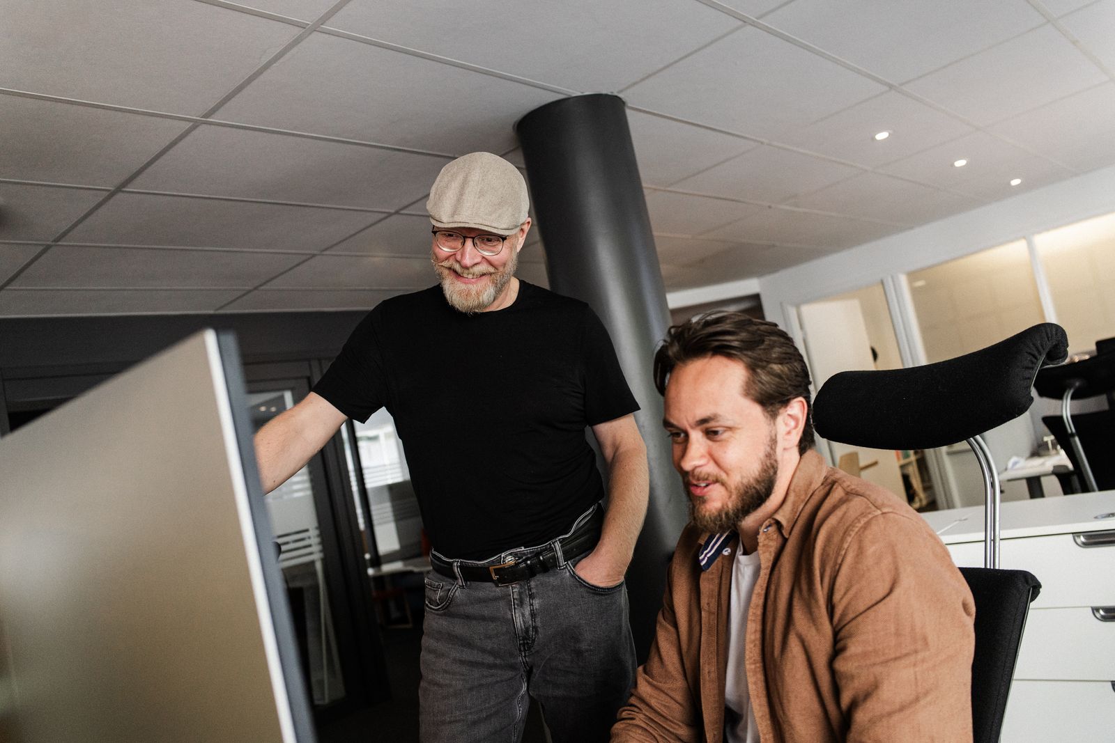 Kim og Mats ser på samme skjerm og smiler. Mats sitter på plassen og Kim står ved siden av.