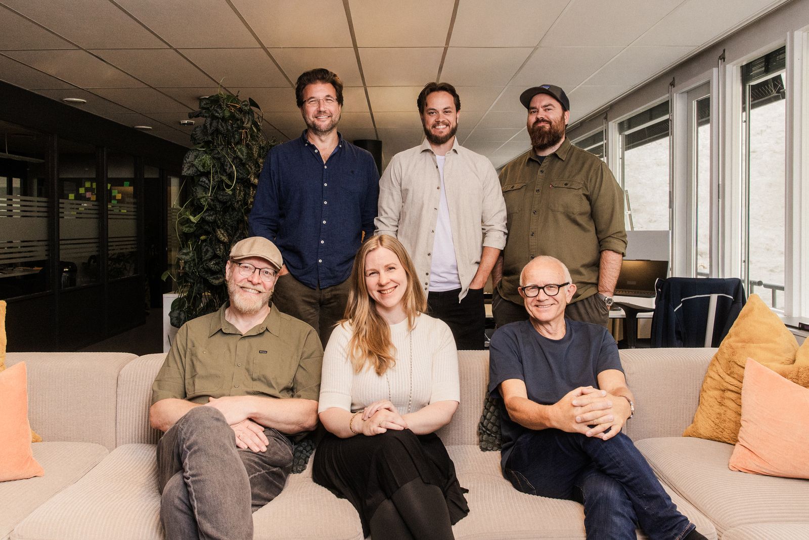 Gruppebilde med alle seks Ravn-ansatte. Tre personer sitter i sofaen, tre personer står bak. Alle ser i kamera og smiler.