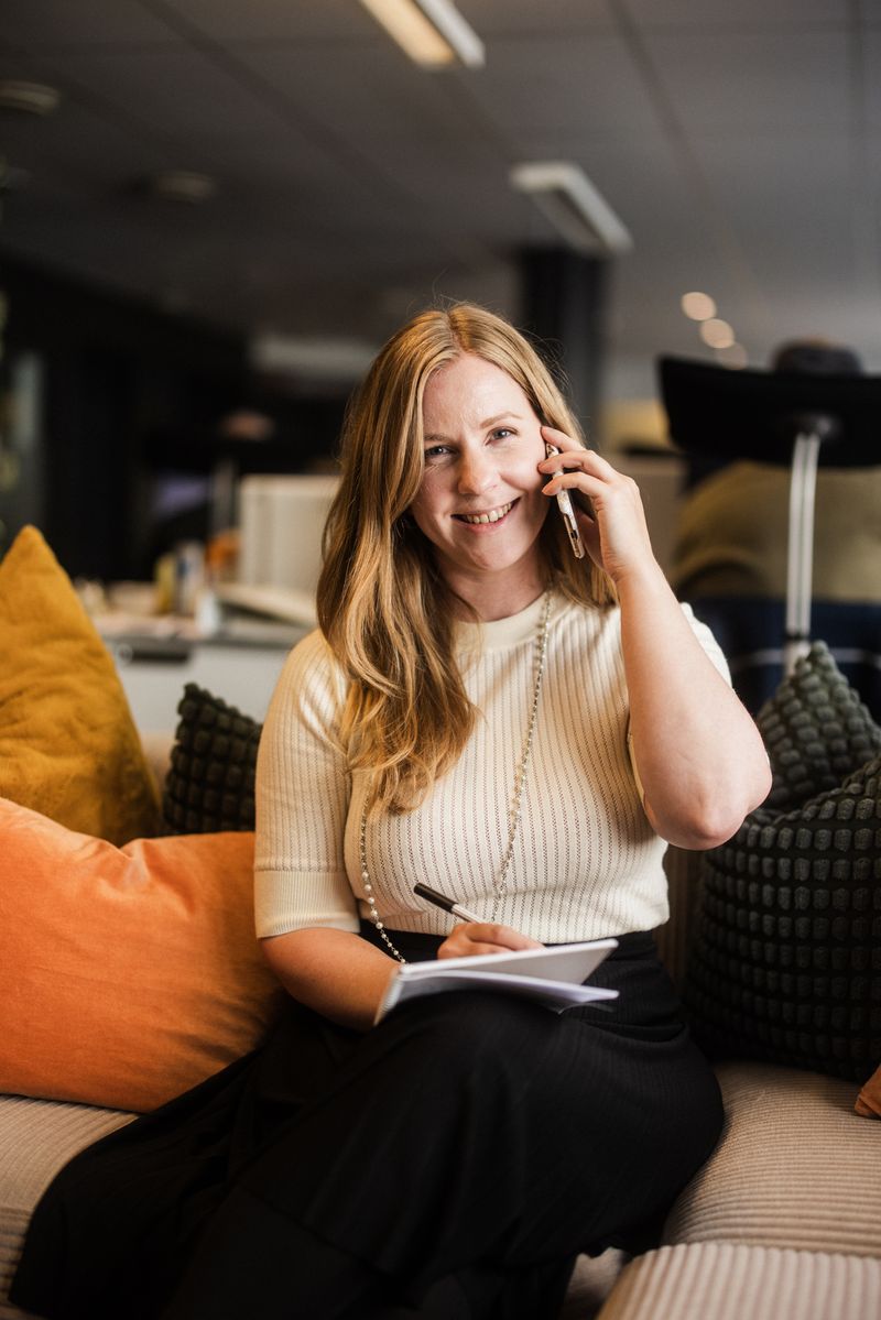 Katrine sitter i sofaen og snakker i telefonen. Hun har penn og skrivebok og ser i kamera mens hun smiler.