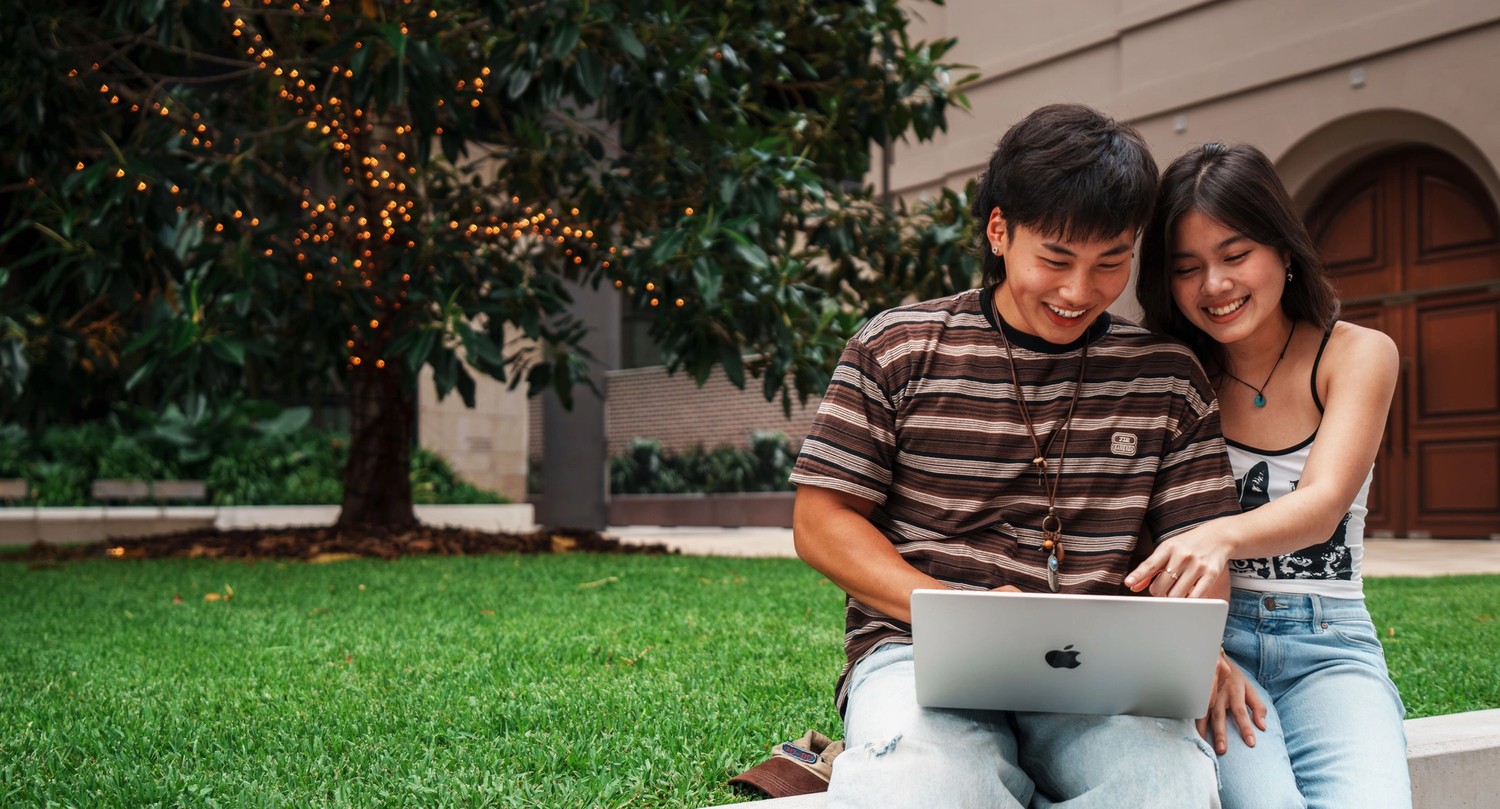 Two young Asian students working on a laptop together in an outdoor green space