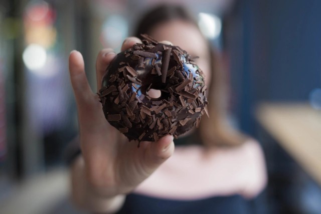 Young female student holding up a chocolate doughnut to the camera
