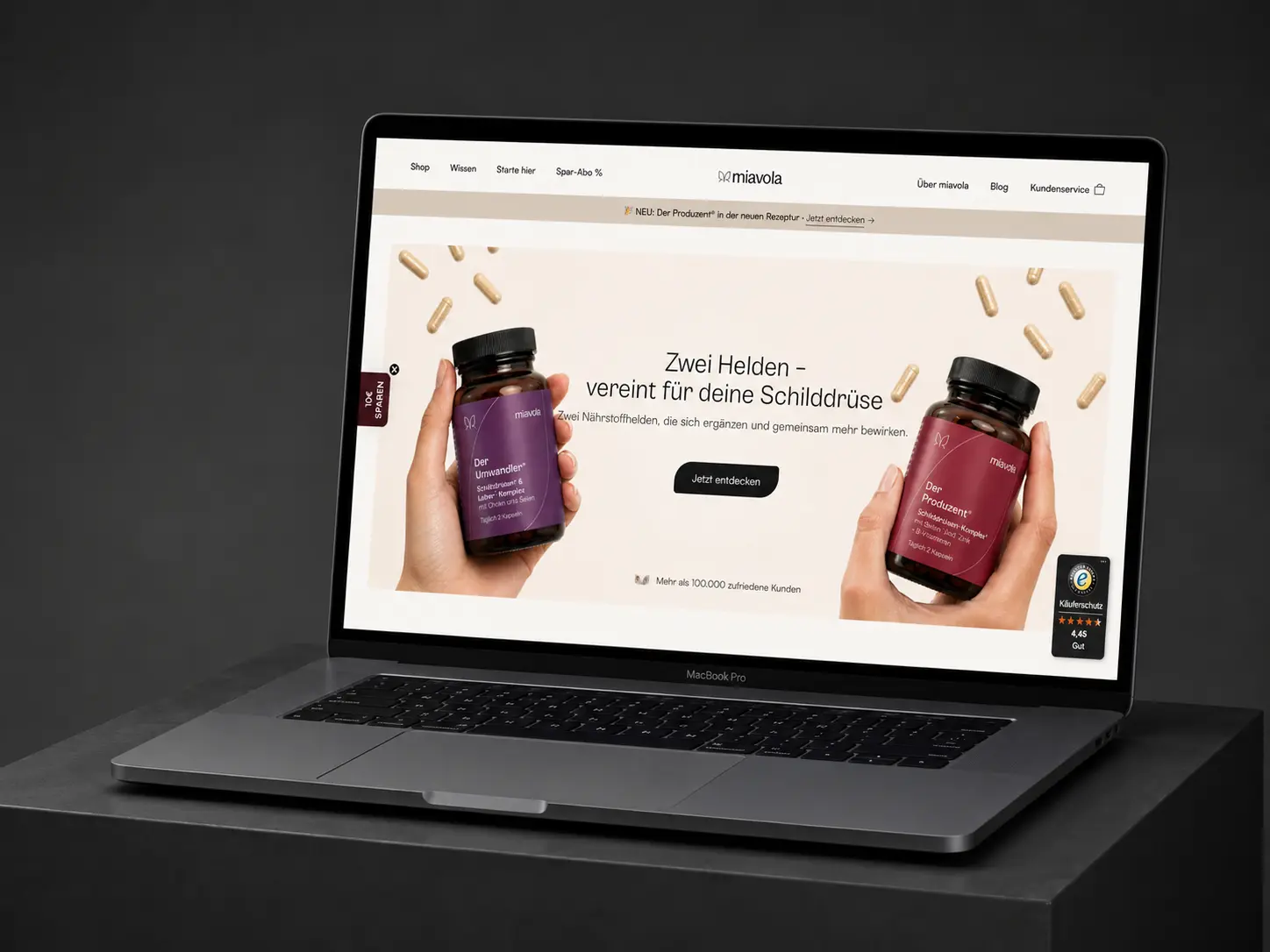 MacBook Pro on a dark plinth showing the miavola homepage: two hands holding the Umwandler and Produzent supplement bottles under the headline "Zwei Helden - vereint für deine Schilddrüse".