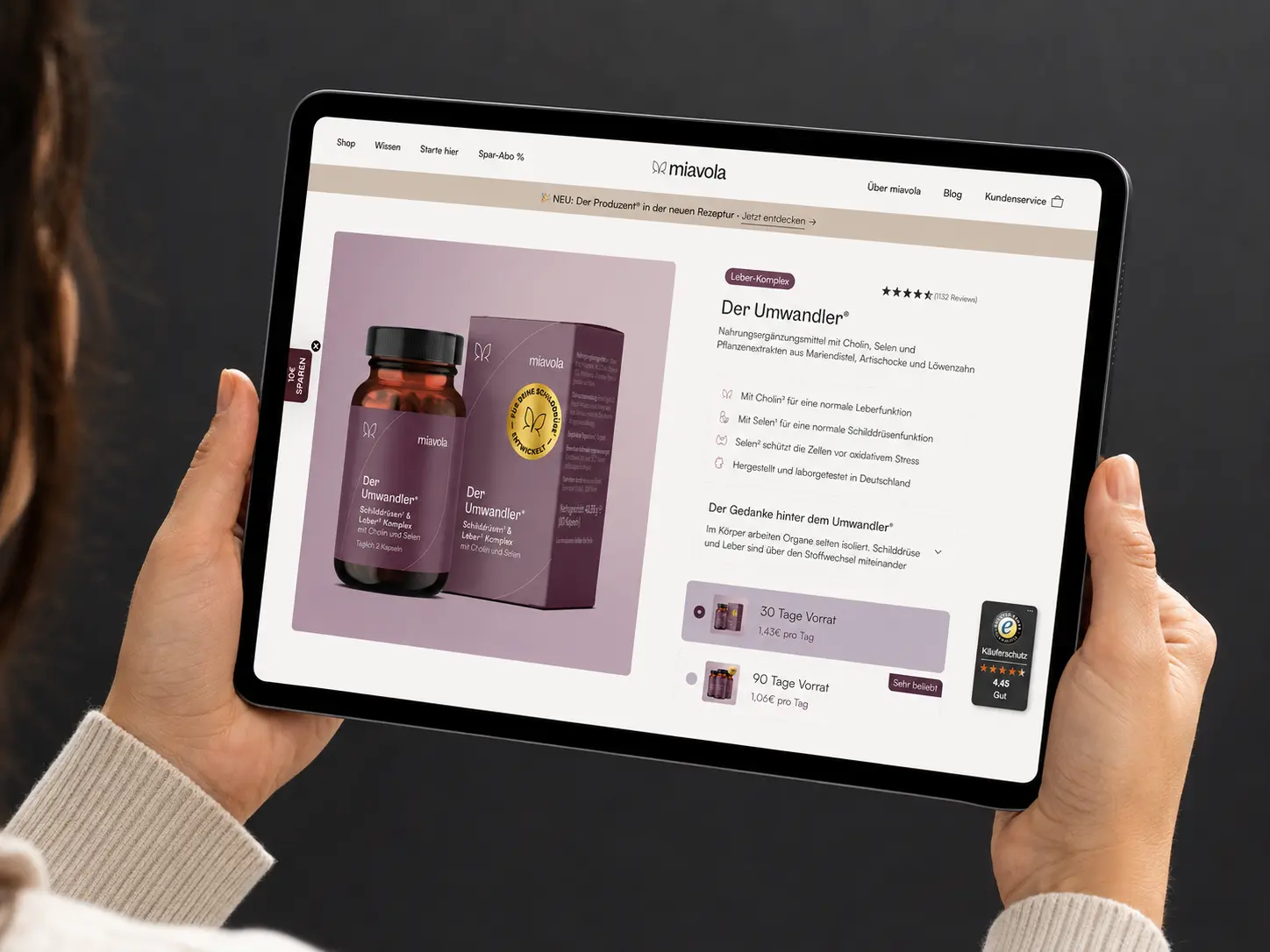 Hands holding a tablet showing the miavola "Der Umwandler" product page, with the bottle and carton, a 4.5-star review score, and 30- or 90-day supply options.