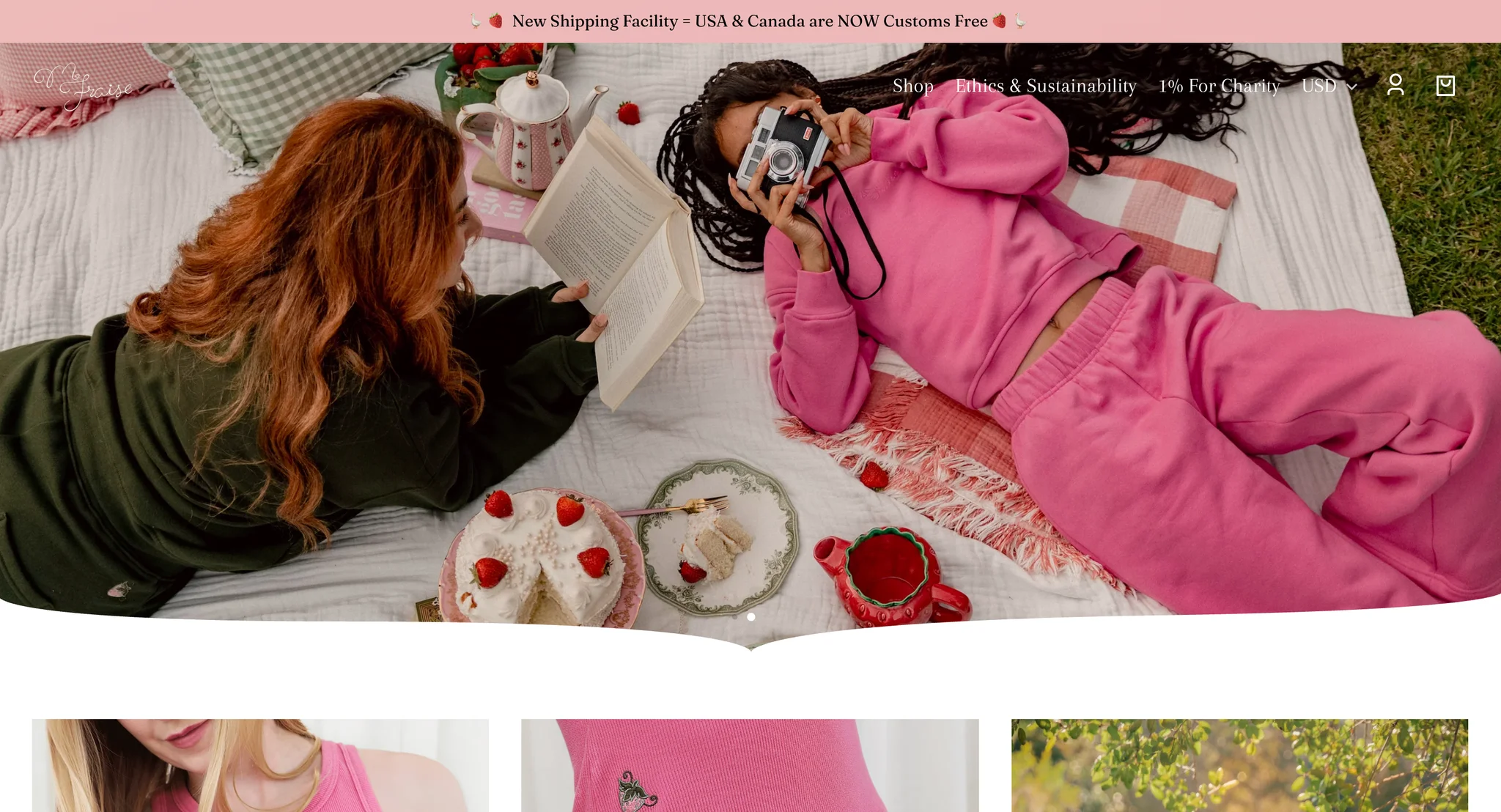 Second homepage slide: two models lying on a picnic blanket beside a strawberry cake and a strawberry-shaped teapot, one in dark green reading a book and one in the pink sweatpants set taking a photo with a film camera