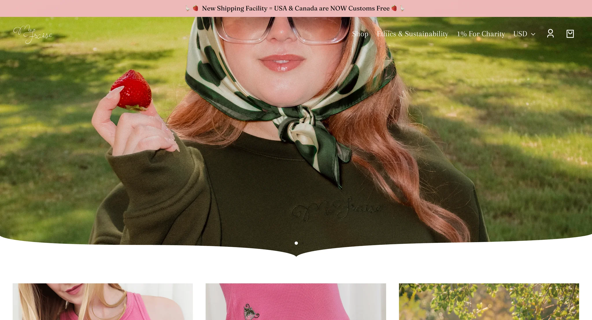 M.Fraise homepage first screen: a pink announcement bar, the white script logo and a menu with Shop, Ethics & Sustainability and 1% For Charity over a full-width photo of a model in a cream and green polka dot headscarf and dark green crewneck holding up a strawberry, with a curved lower edge