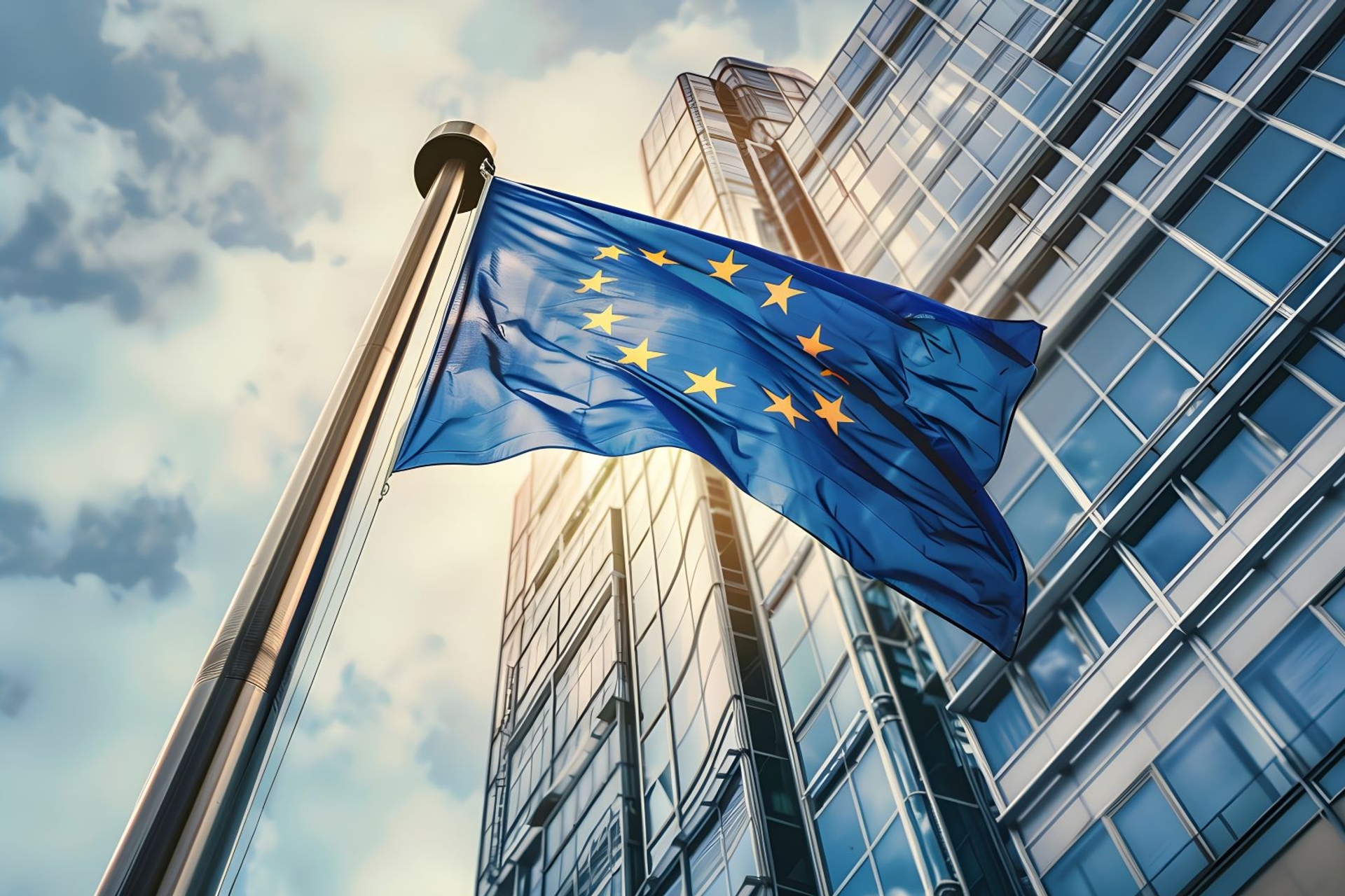 Europese vlag wappert voor een hoog kantoorgebouw met veel glas en blauwe lucht met wolken.